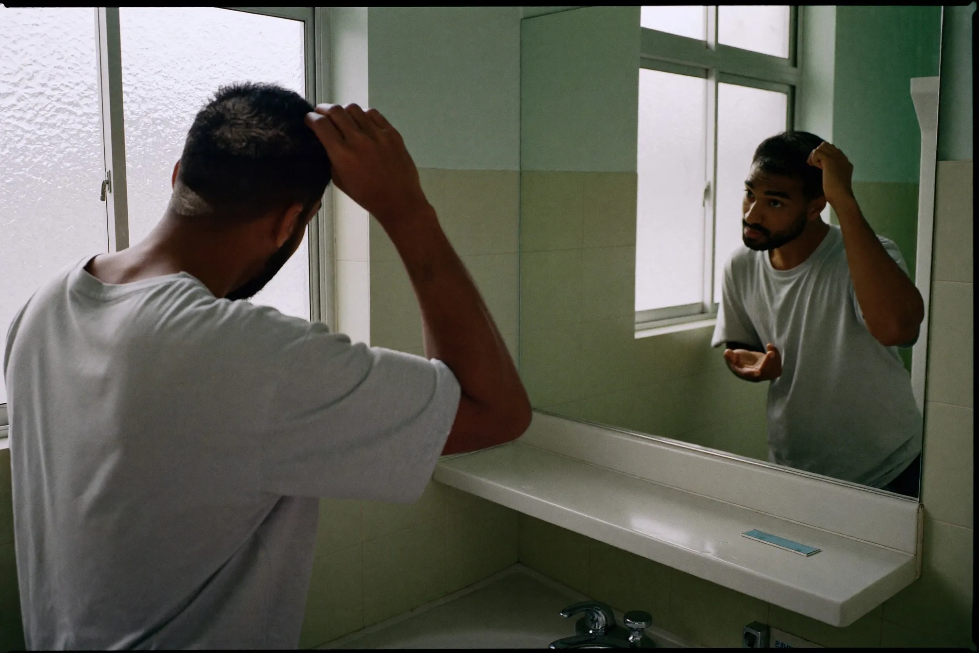 Indian Singaporean man pushing his fringe forward with a fingertip of matte clay in front of an HDB bathroom mirror