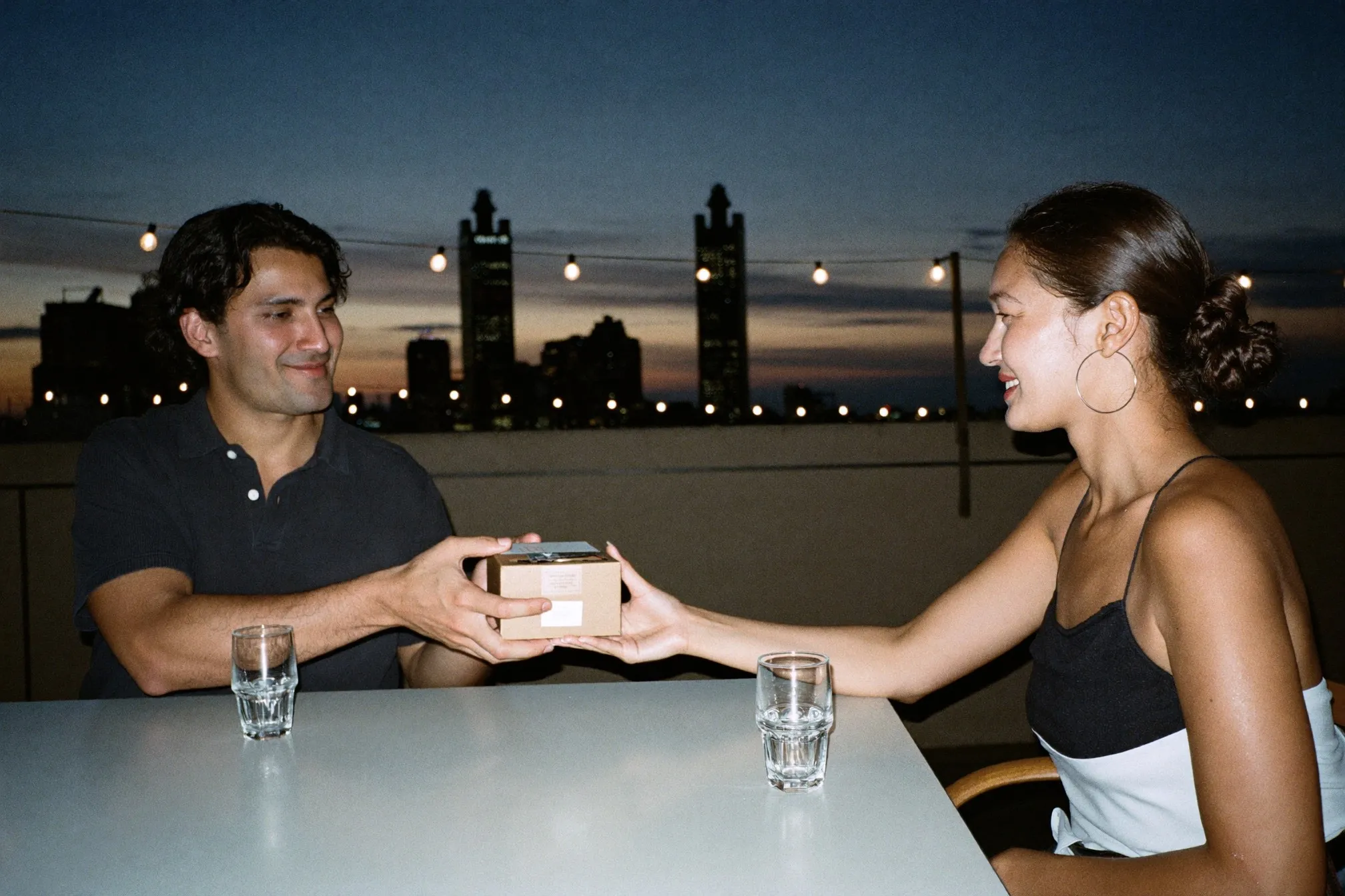 A man passing a small wrapped box across a table at a rooftop bar in Kuala Lumpur at dusk