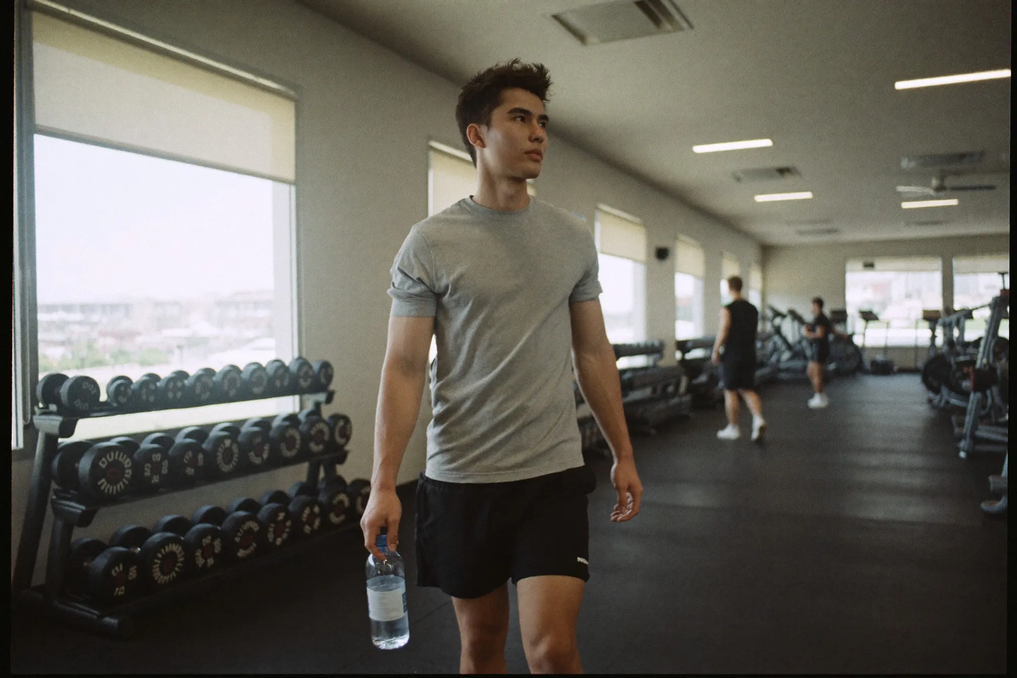 Young Malay man in a grey t-shirt walking between dumbbell racks in a bright KL gym at mid-morning