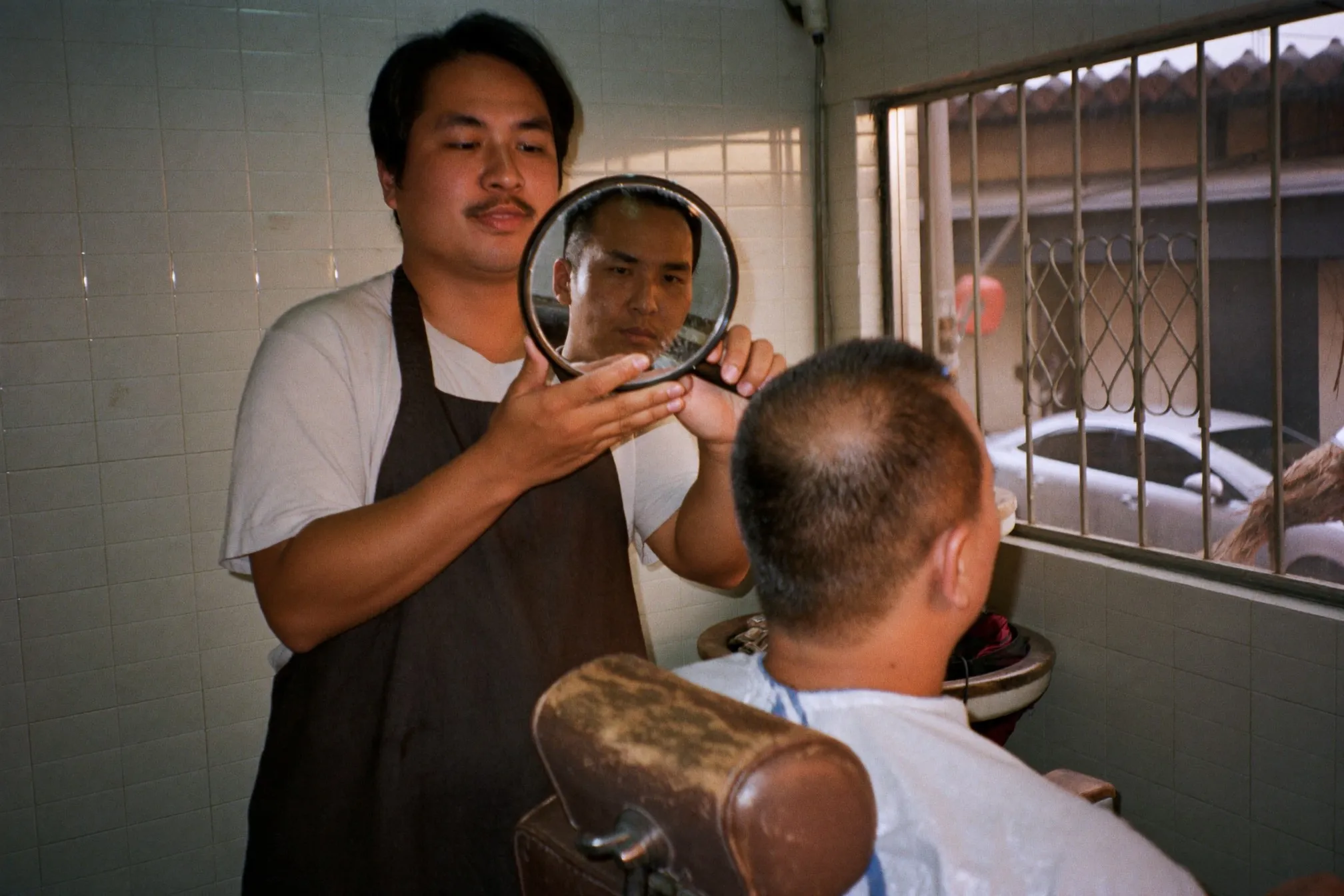 Barber holding a hand mirror behind a young man in the chair so he can see the crown of his head
