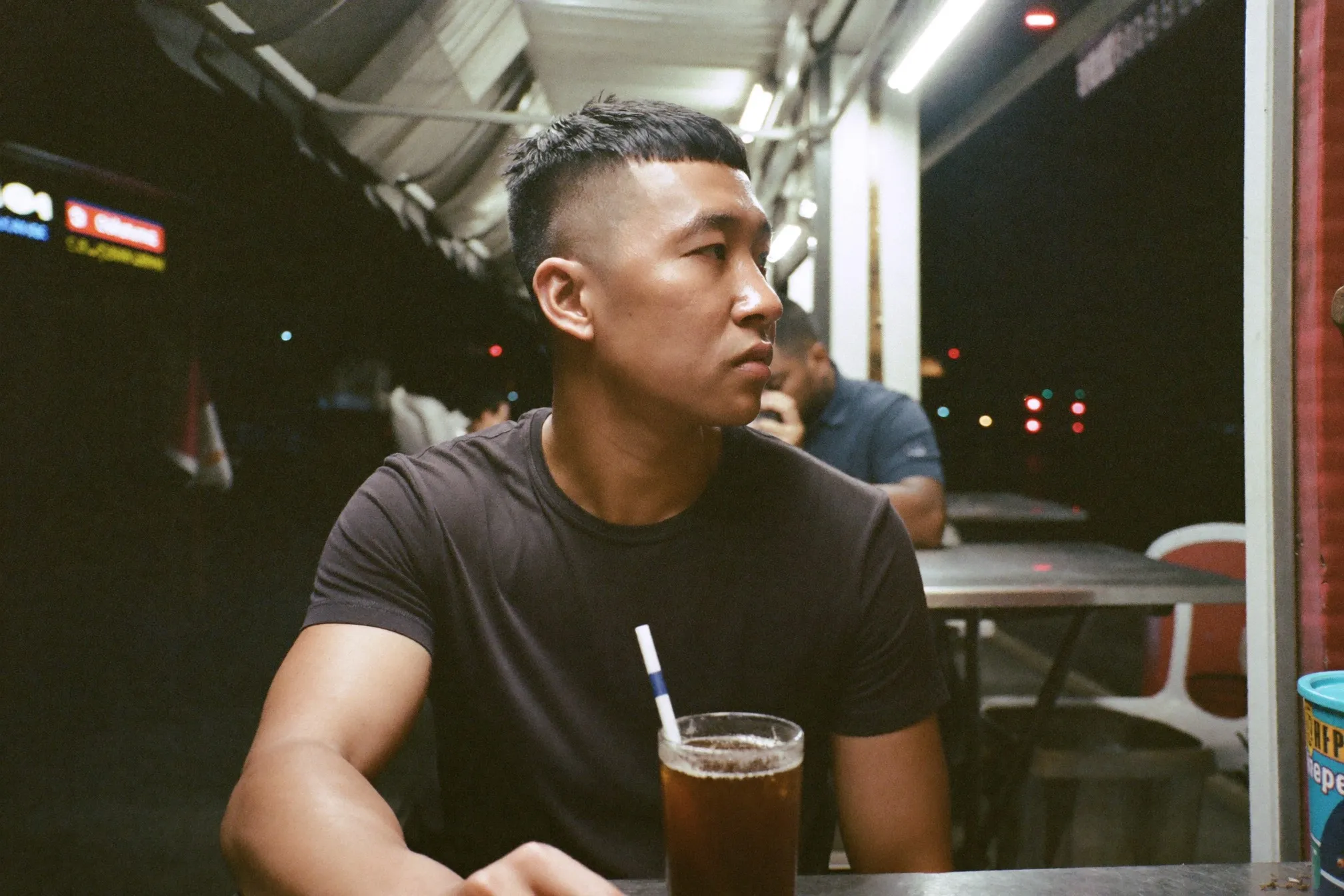 Man with a matte textured crop sitting at an open-air mamak table on a humid Kuala Lumpur evening