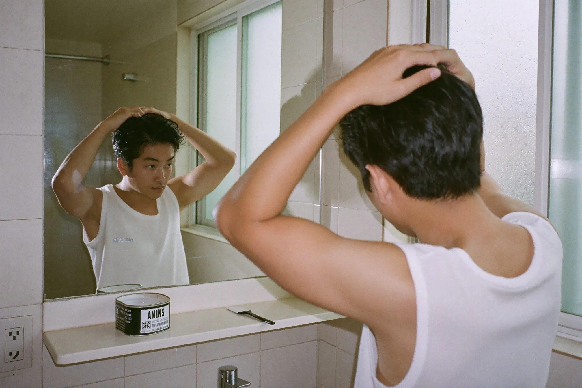 Man styling his hair with pomade in front of a bathroom mirror in an HDB flat, morning light through the window