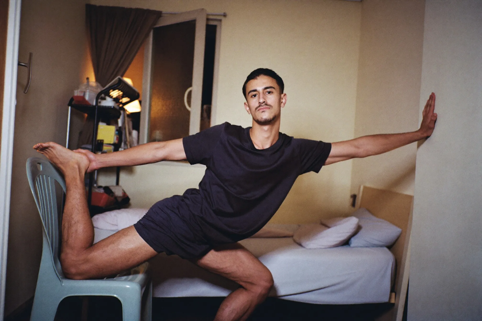 Malay man doing a rear-foot-elevated split squat with his back foot on a plastic chair in a narrow HDB bedroom