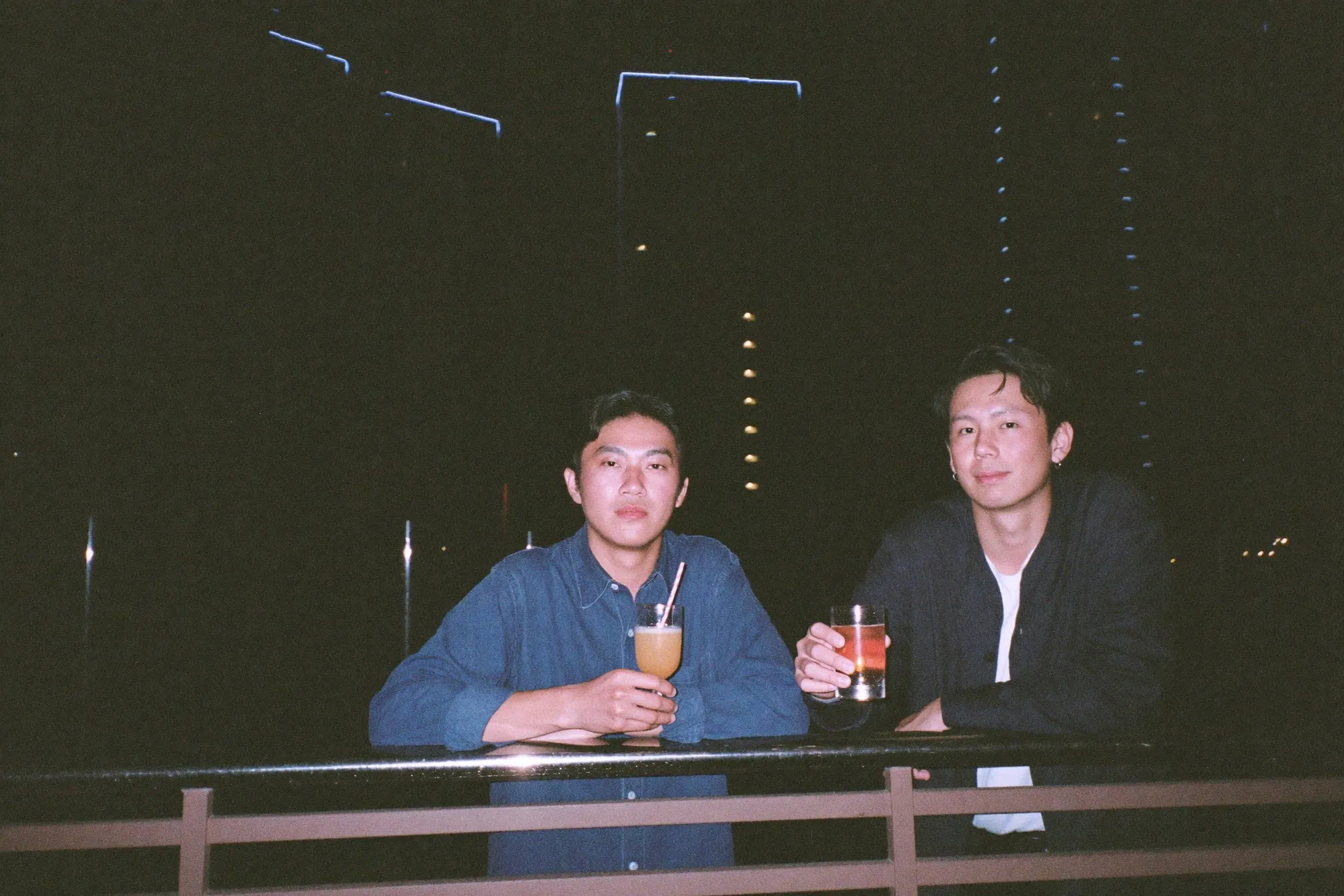 Two men leaning on a condo balcony railing at night with drinks, KL apartment towers lit behind them