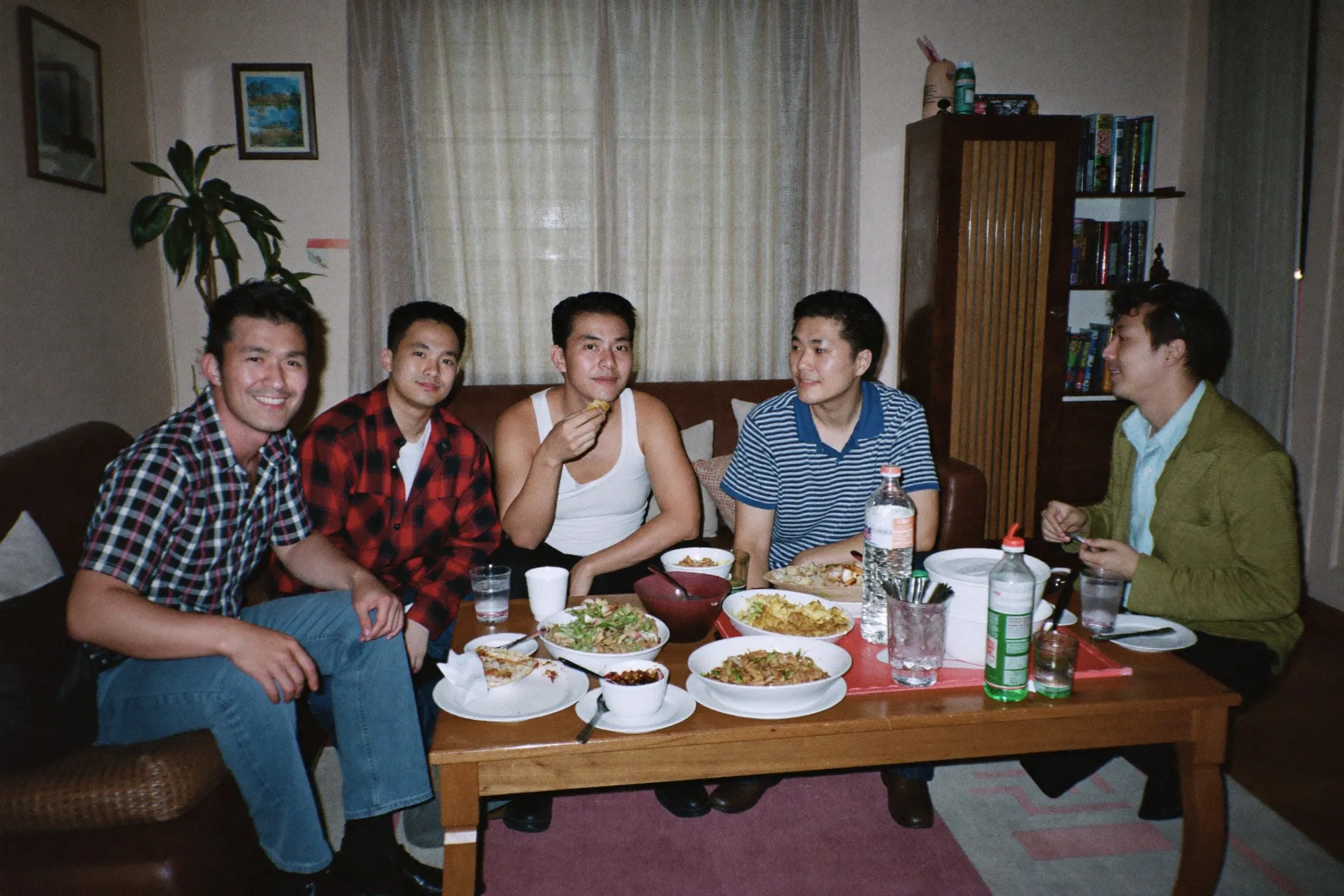 Six friends sitting and standing around a low coffee table of shared food in a small HDB living room in the evening