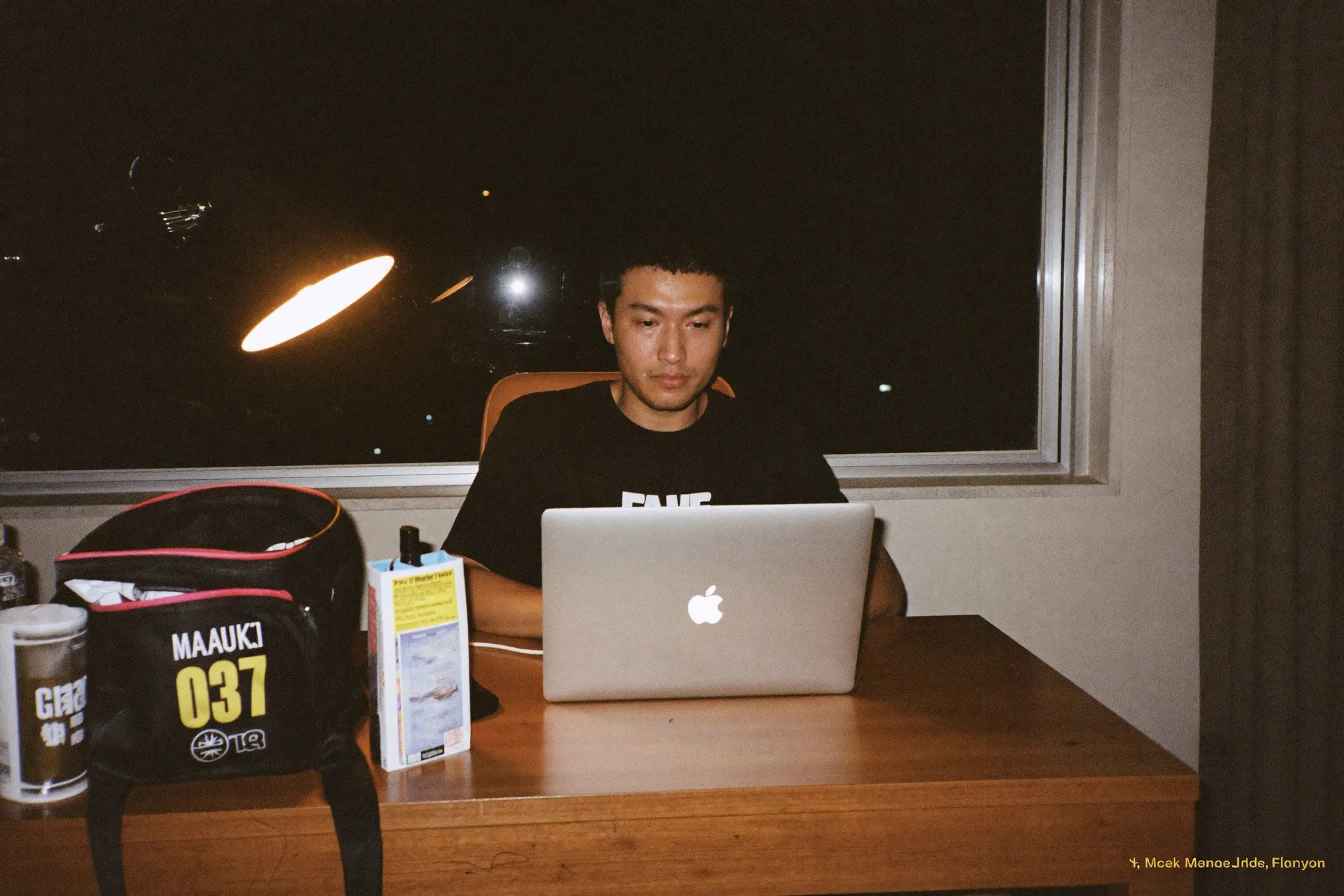 Man working on a laptop at a tidy desk at night, warm lamp, gym bag by the chair