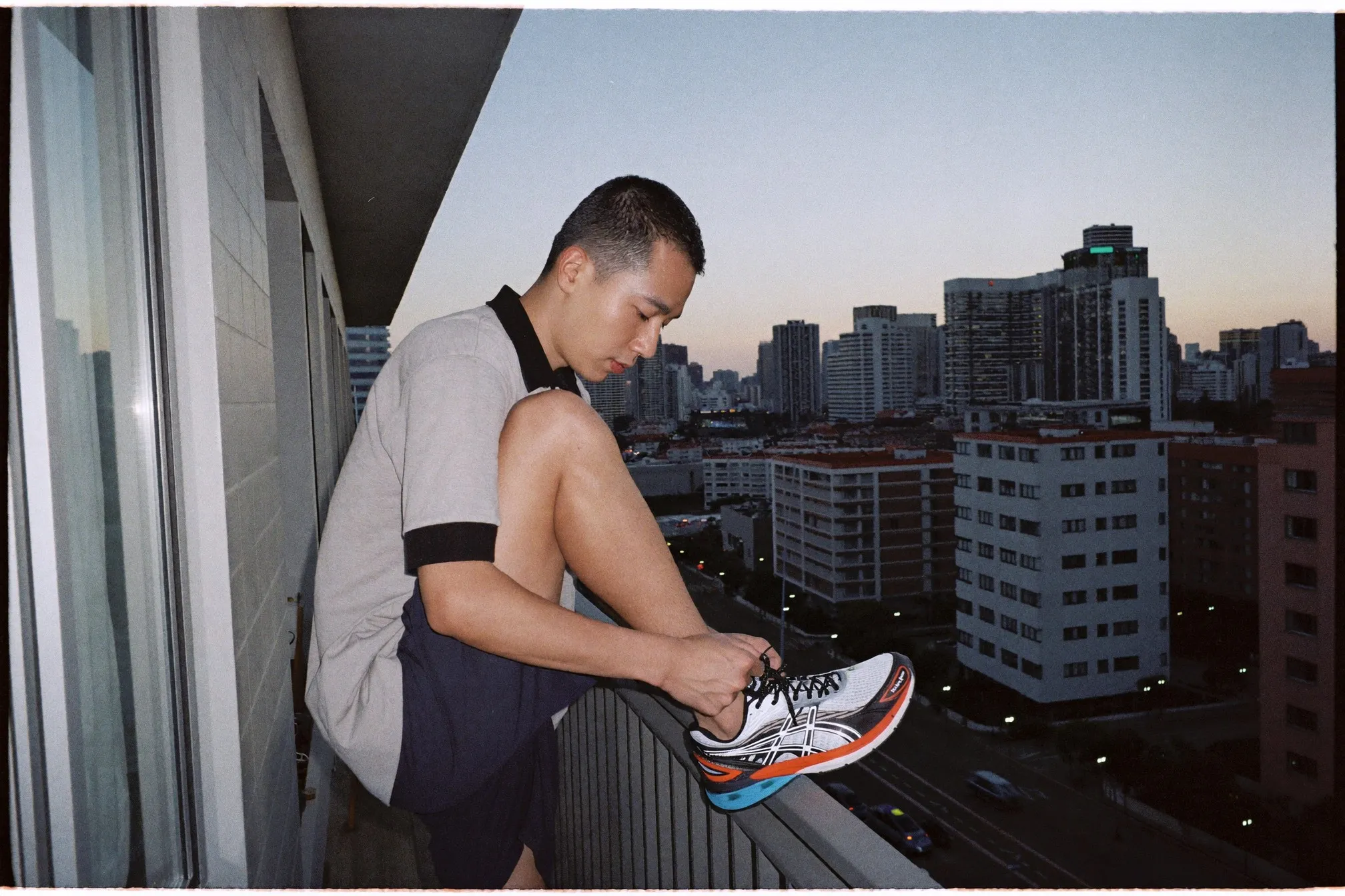 Man lacing running shoes at dawn on an apartment balcony, city skyline behind him
