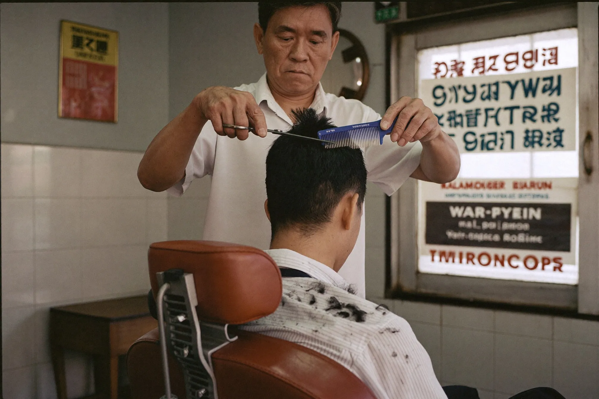 Barber scissor-cutting the top of a Malay man's ivy league haircut over a comb in a small Kuala Lumpur barbershop
