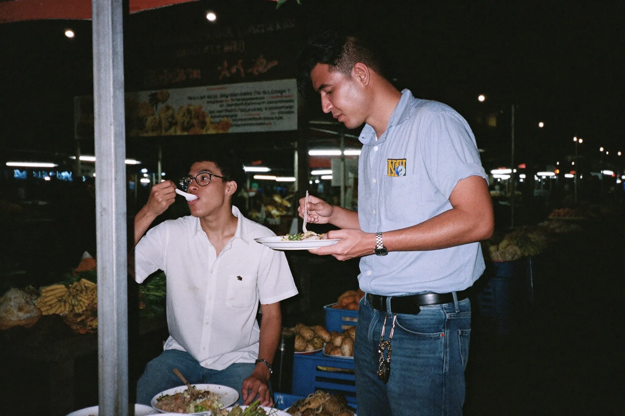Two friends in jeans and short-sleeve shirts eating at an open-air hawker centre at night