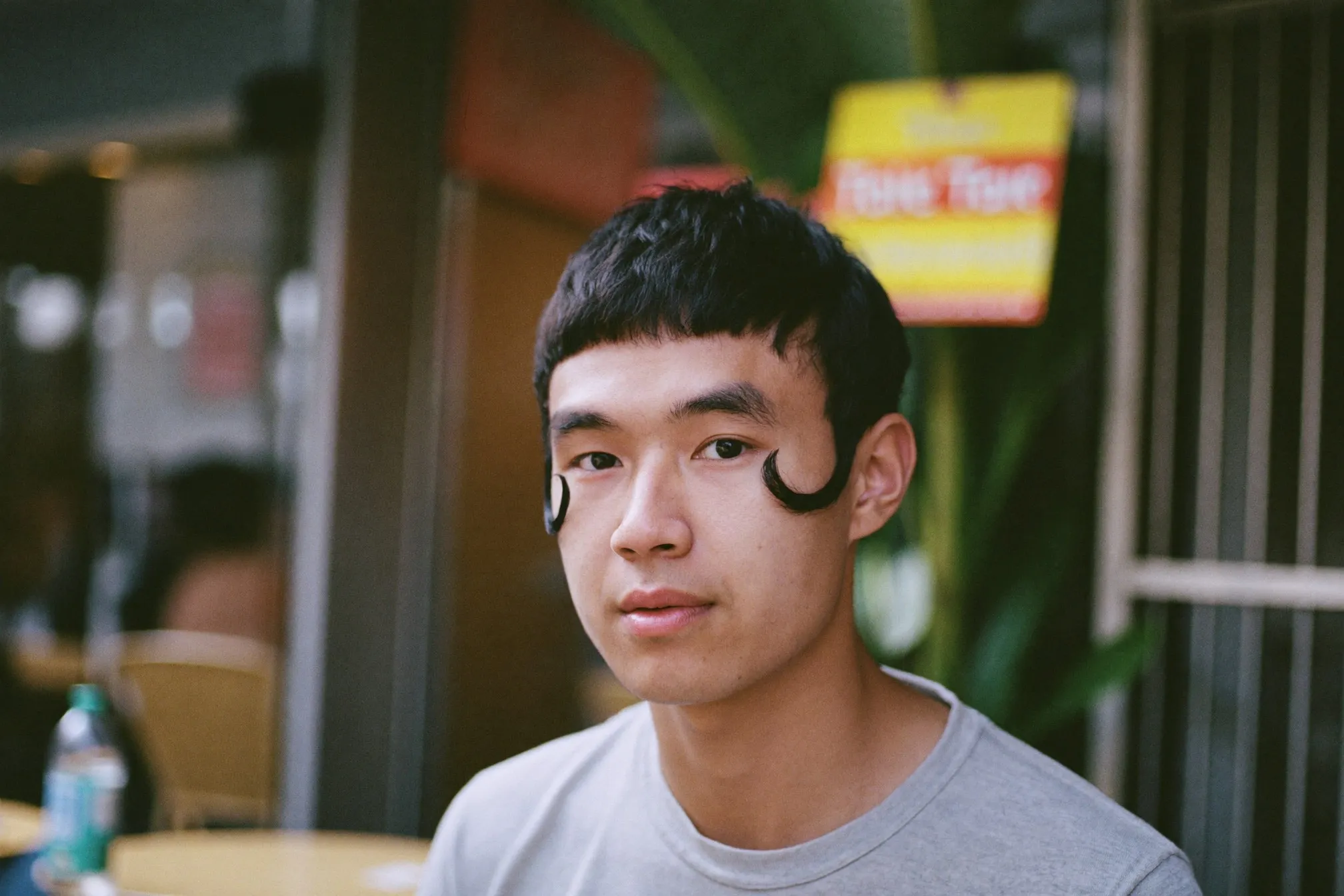Close portrait of a young Malay man with comma hair, the fringe curling inward at his temples, outside a cafe in Bangsar