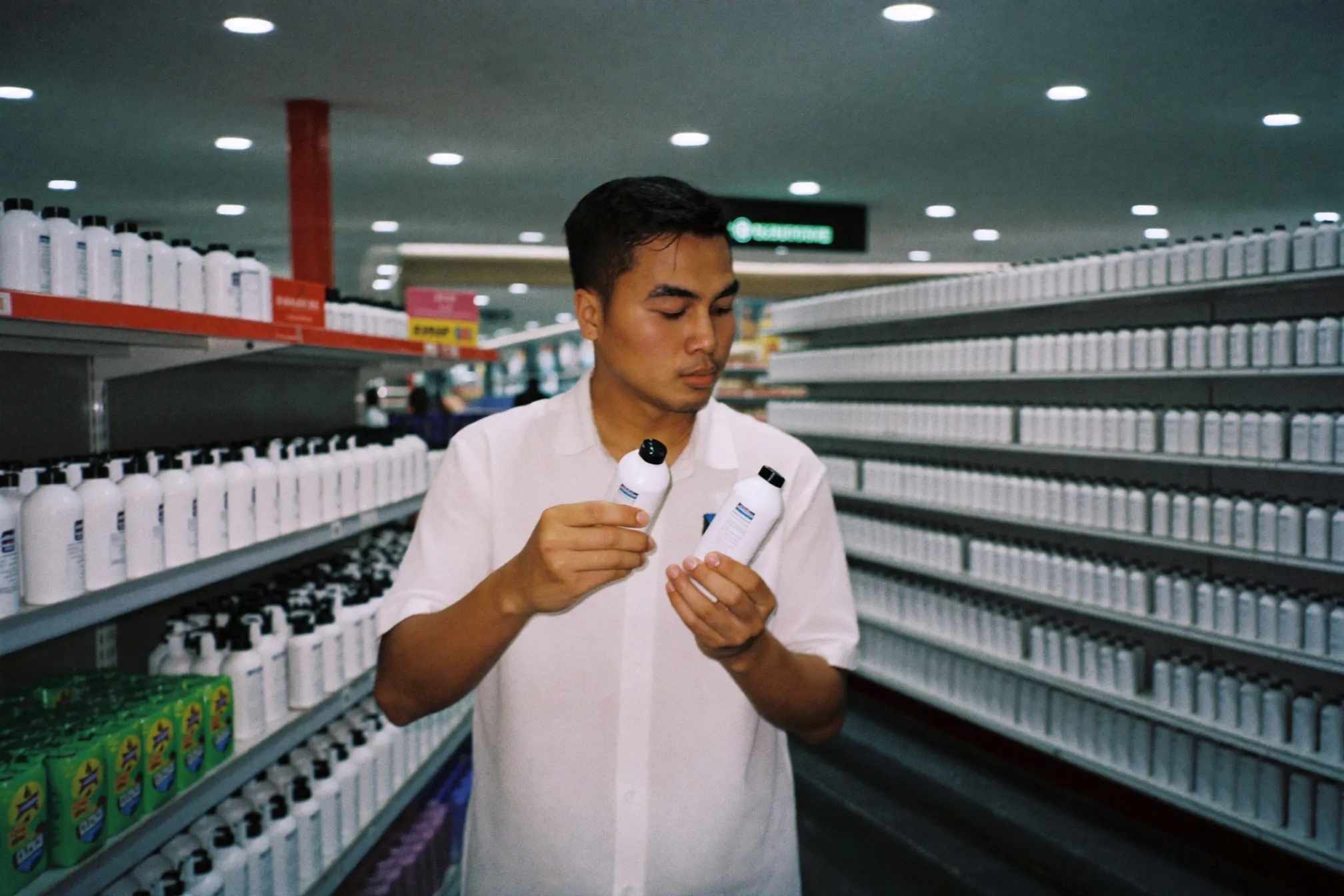 Man comparing two plain unbranded skincare bottles in a brightly lit pharmacy aisle