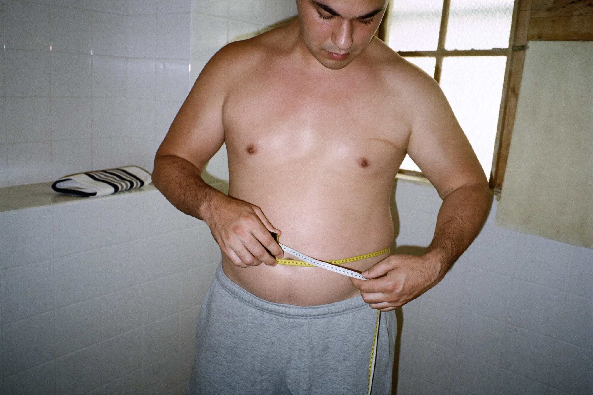 Close view of a man measuring his waist with a soft tape measure at navel height in a bathroom