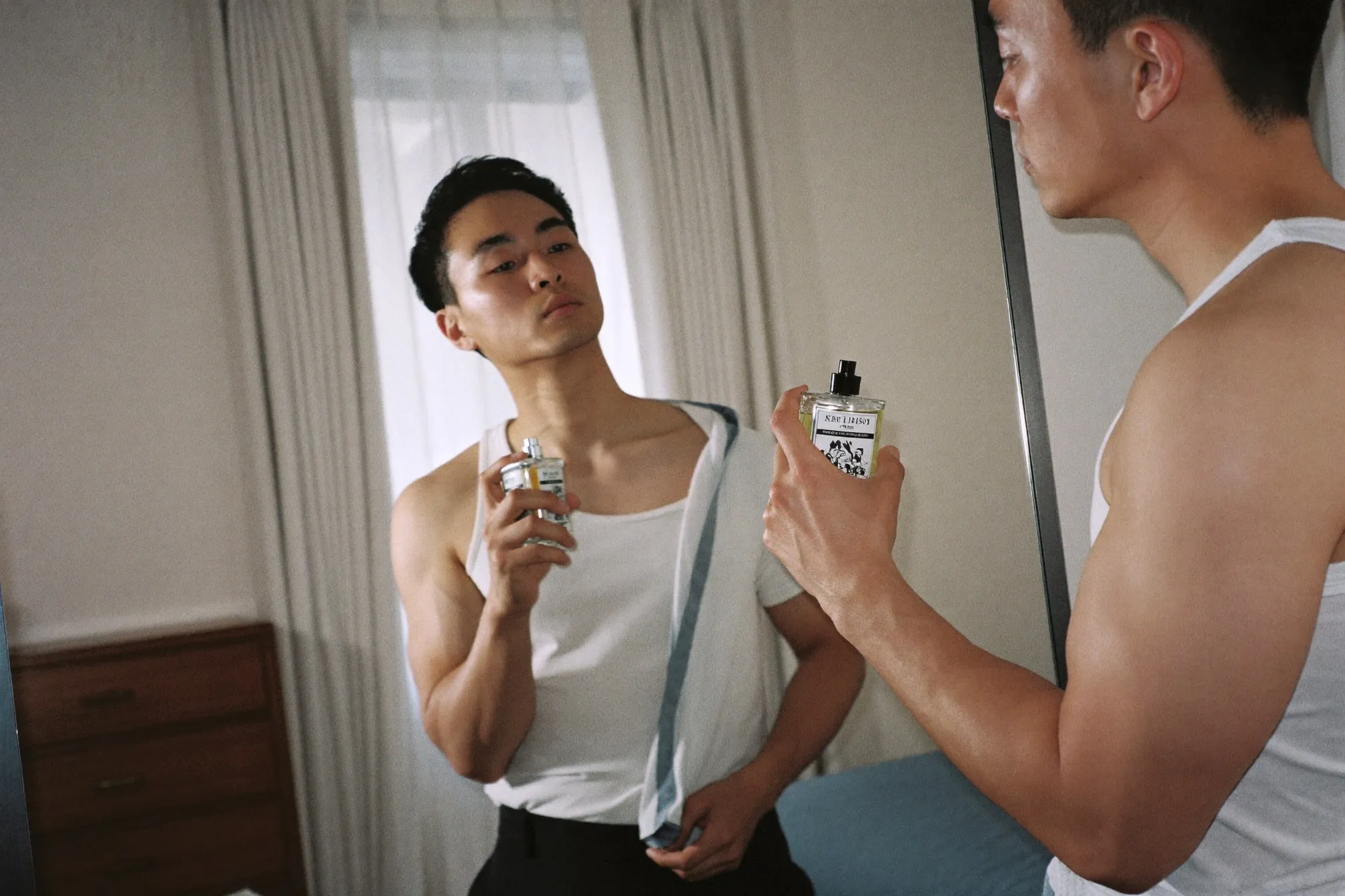 Man in an undershirt spraying fragrance onto his chest in front of a bedroom mirror in morning light