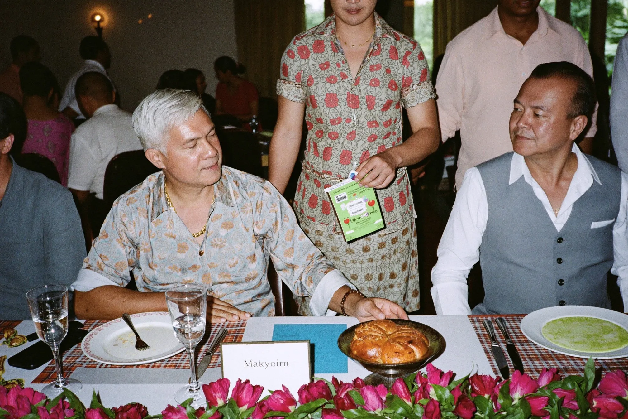 Man in a batik shirt handing a green packet to a groom at a wedding reception table