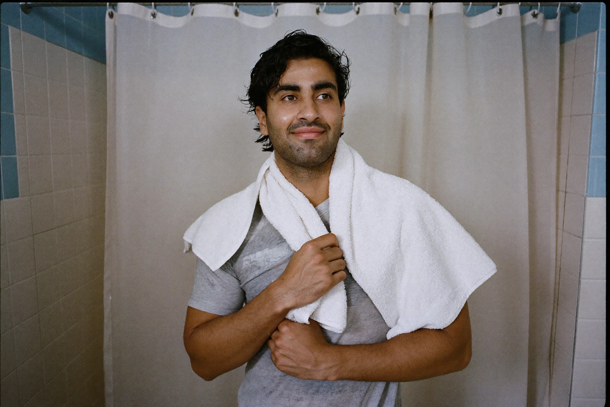 Man with a towel over his shoulders standing in a tiled flat bathroom after a shower