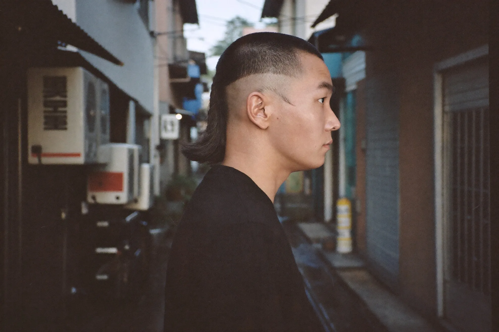 Three-quarter rear view of a young Chinese Singaporean man with a burst fade mullet standing in a shophouse alley at dusk