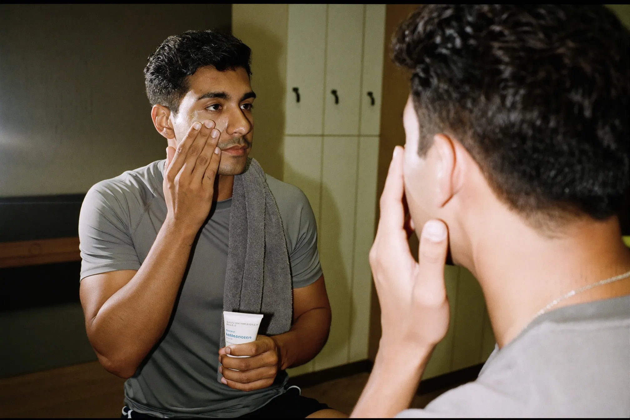 Man applying moisturiser at a gym locker room mirror after a workout, towel over his shoulder