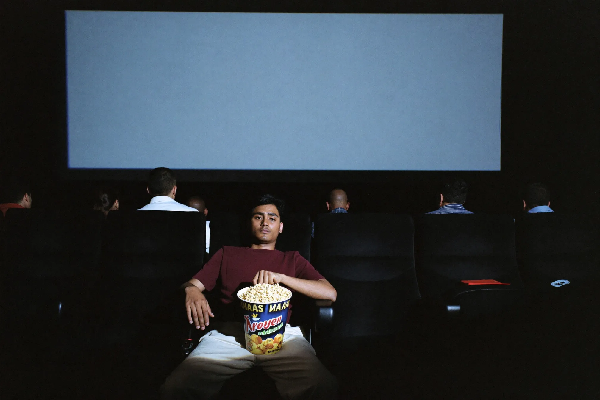 Cinema auditorium seen from the back row, audience silhouettes against the glow of a wide screen