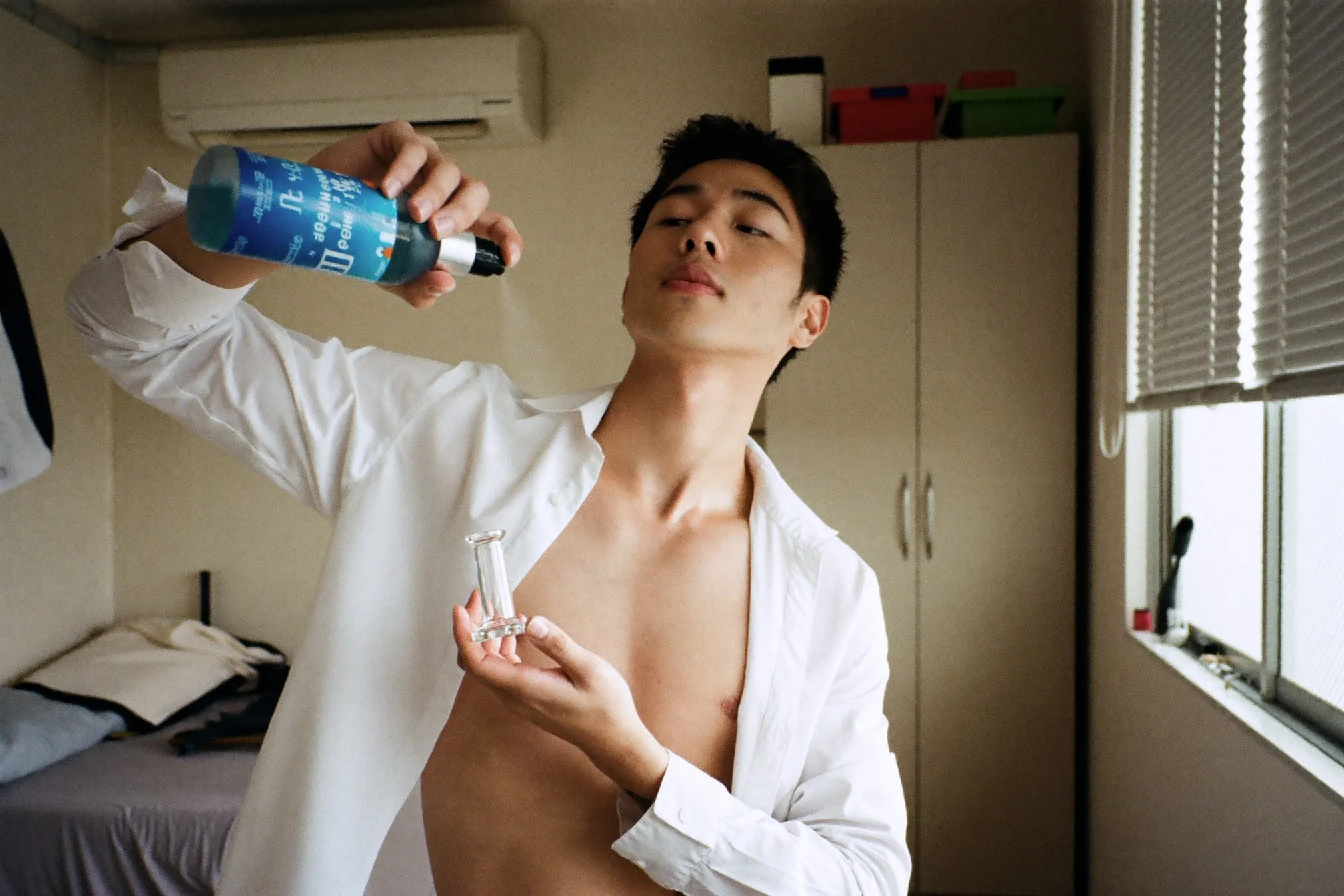 Man spraying an unbranded frosted glass bottle onto his chest under an open shirt in an HDB bedroom before work