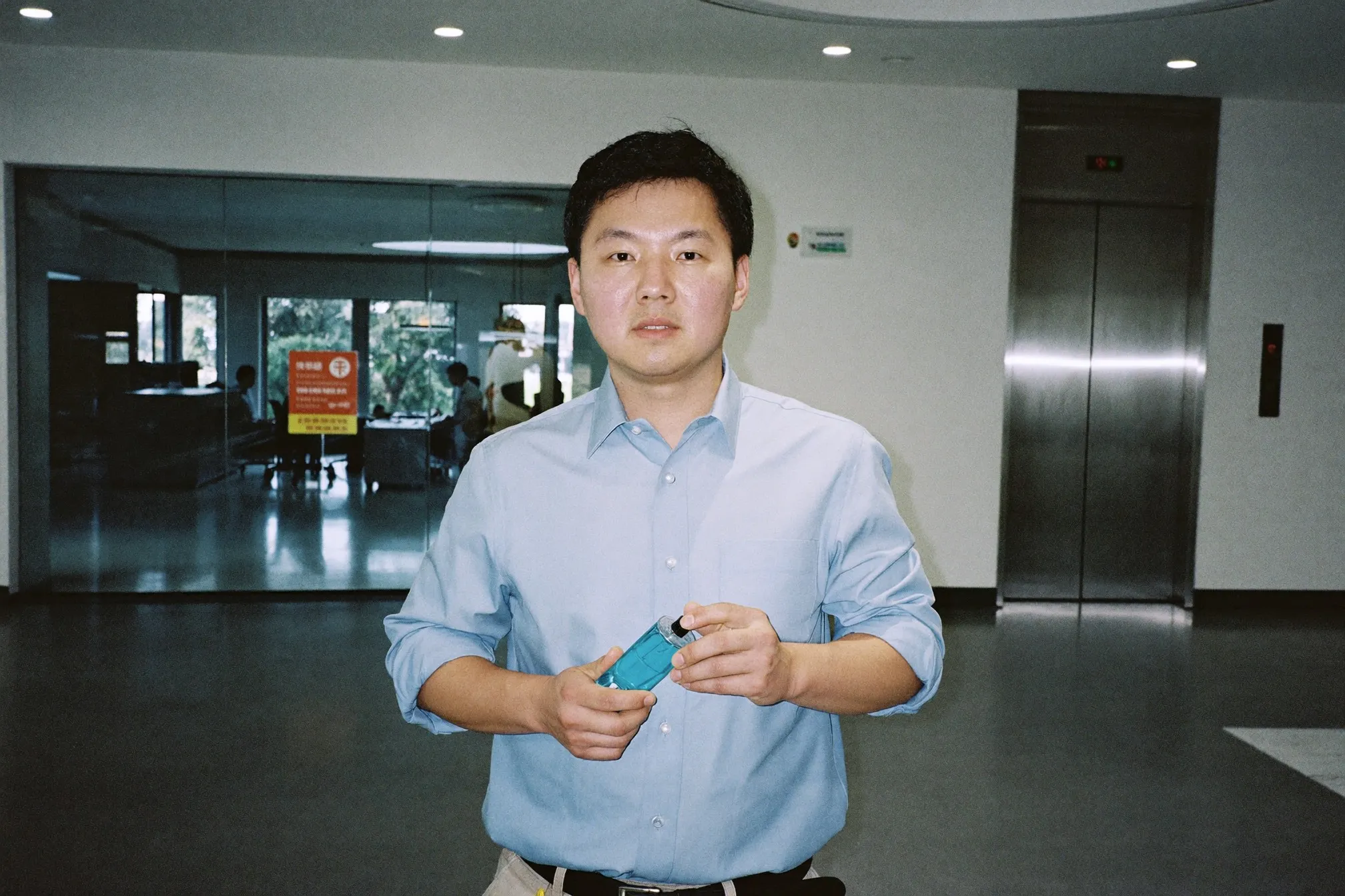 Man in a pale blue office shirt holding a small unbranded glass atomiser in a Kuala Lumpur office lift lobby in the morning