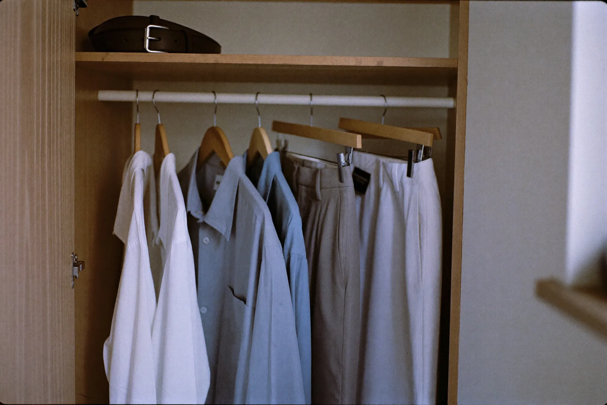 Work shirts in white, light blue and grey hanging beside pressed trousers and a leather belt in a bedroom wardrobe