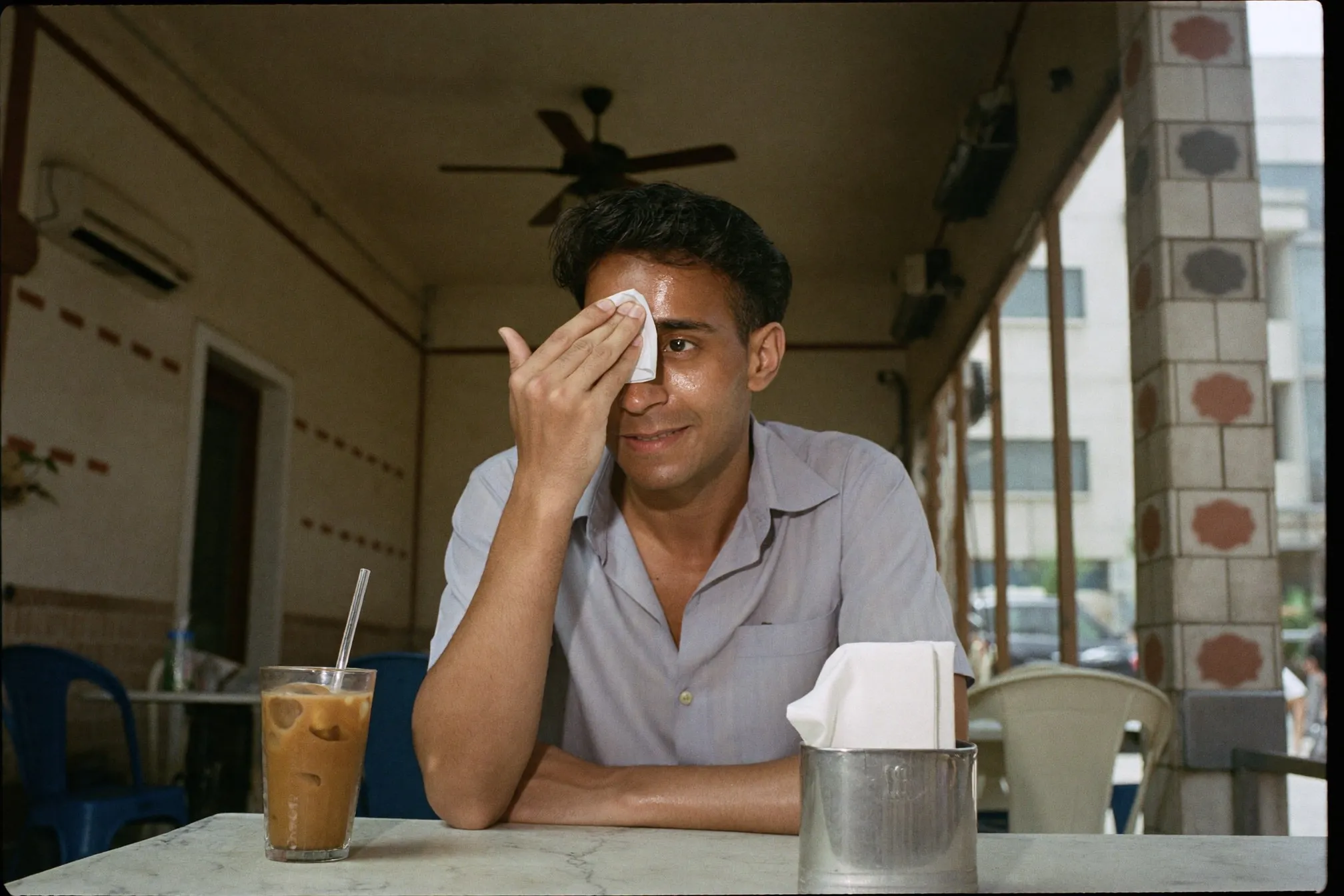 Man blotting shine from his forehead with a tissue at an outdoor kopitiam in the afternoon heat