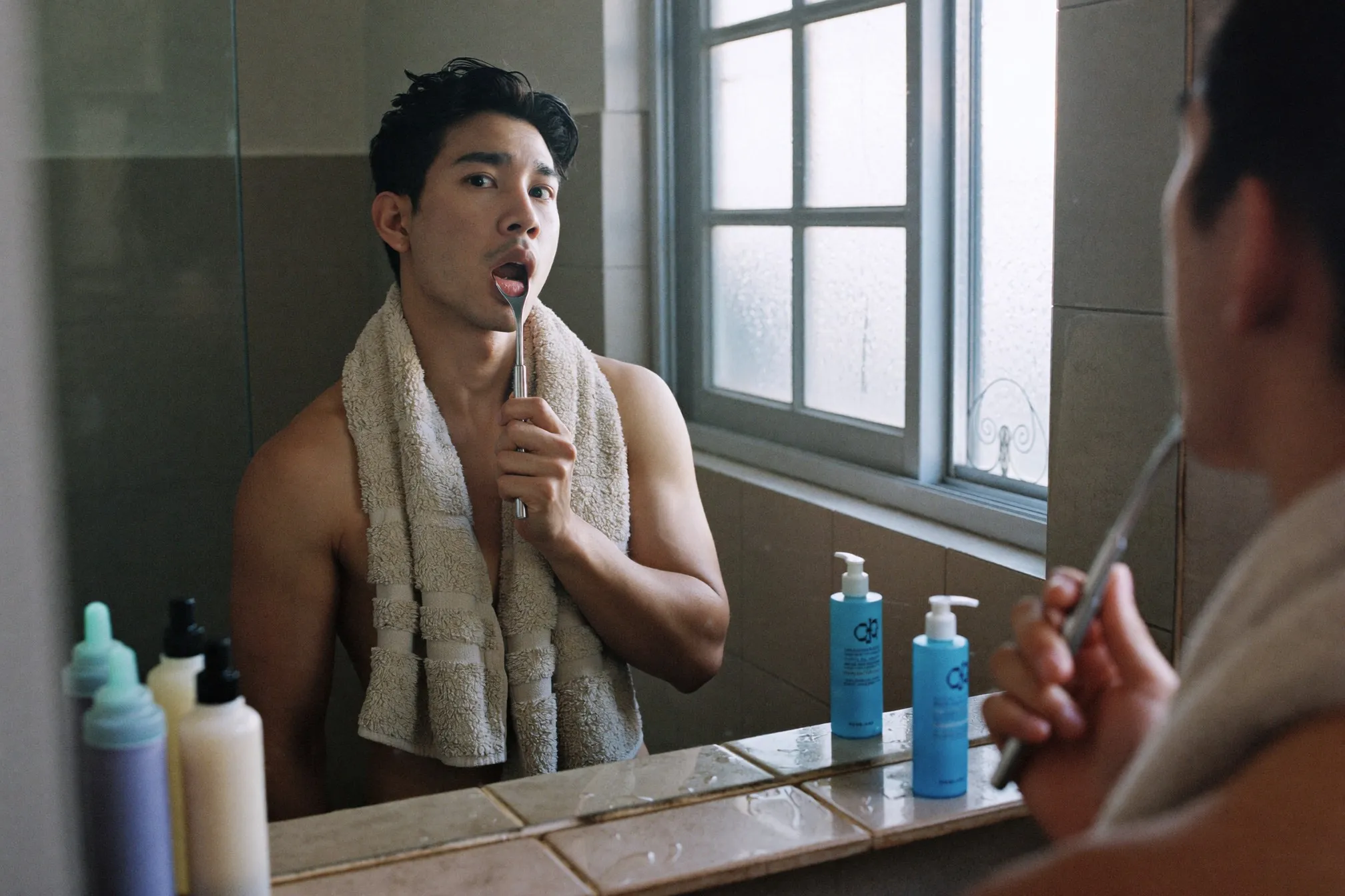 Man at a bathroom mirror in the morning holding a plain metal tongue scraper