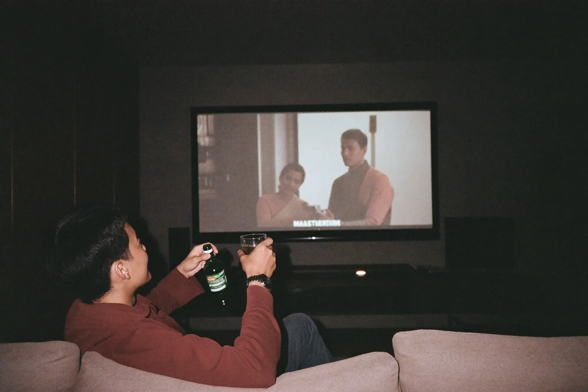 Man settling into a sofa with a drink as a home cinema screen glows in a dark living room