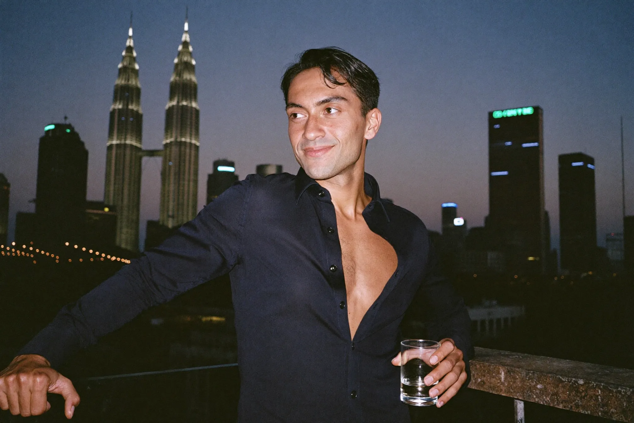 Man in an open-collar shirt at a rooftop bar in the evening, city skyline behind him