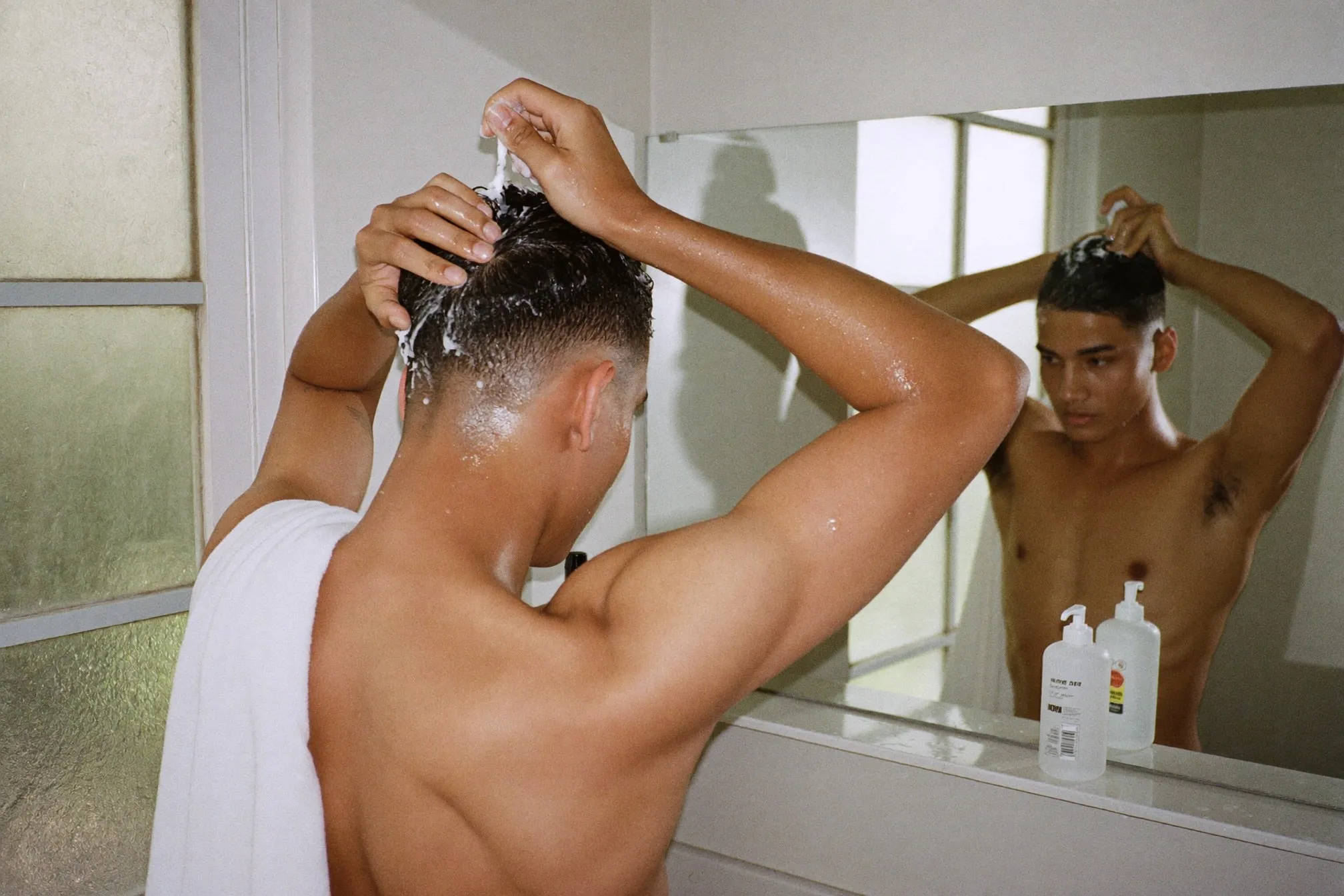 Man with a grown-in permed fringe scrunching curl cream into damp hair in a bathroom mirror