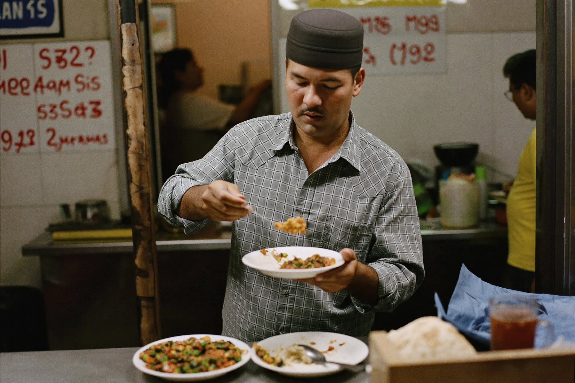 Indian Malaysian man ordering at a late-night mamak stall in Kuala Lumpur with roti and eggs on the counter