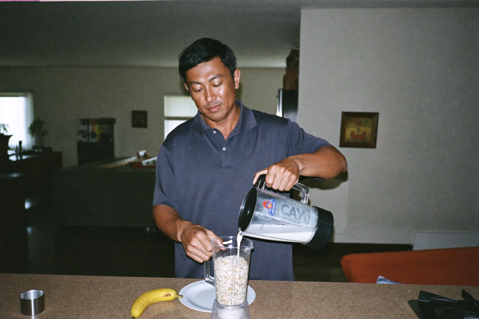 Chinese Malaysian man pouring milk into a blender jug on a kitchen counter in a KL condo, banana and oats beside him