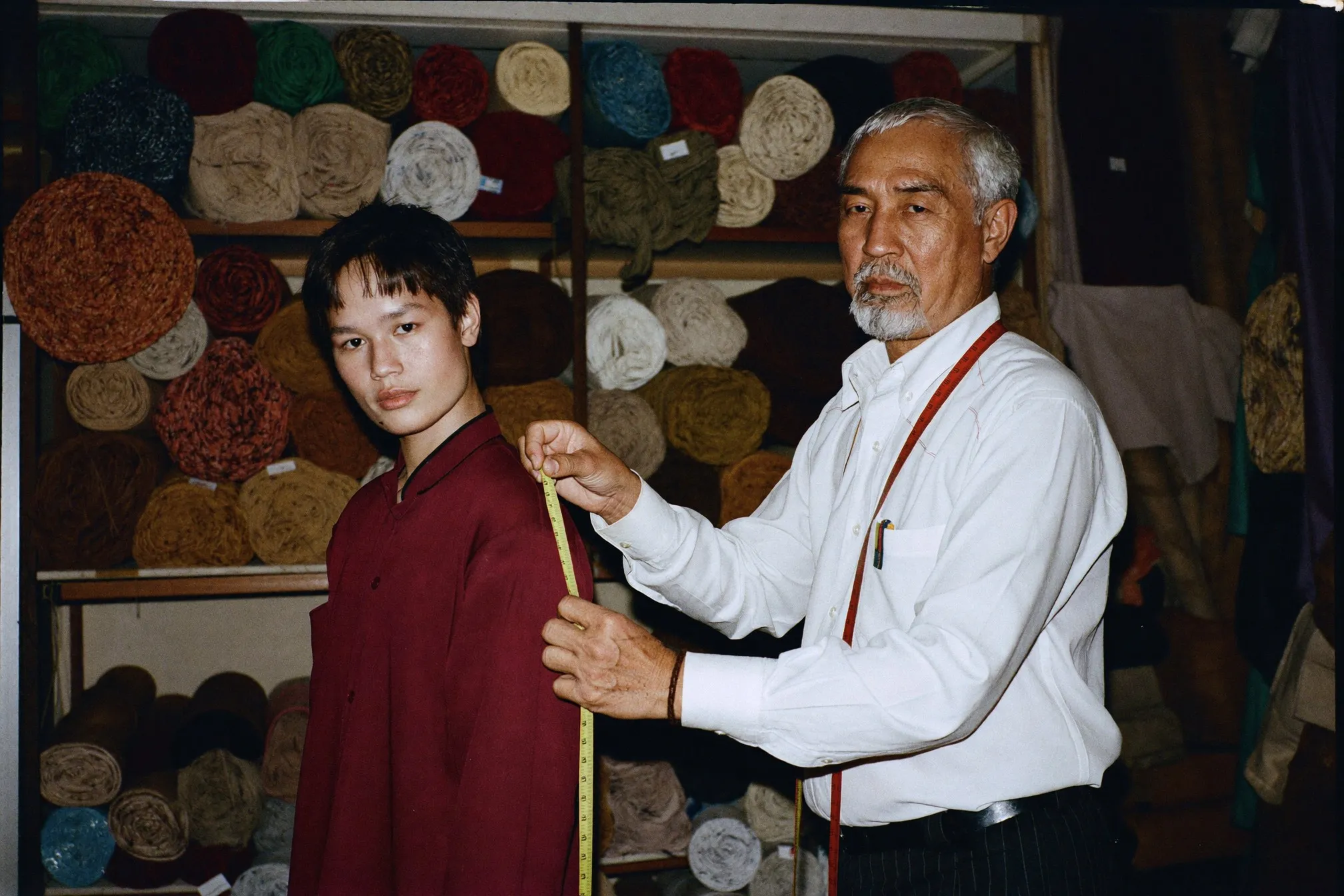 Tailor measuring the shoulder of a young man for a baju melayu inside a small fabric shop