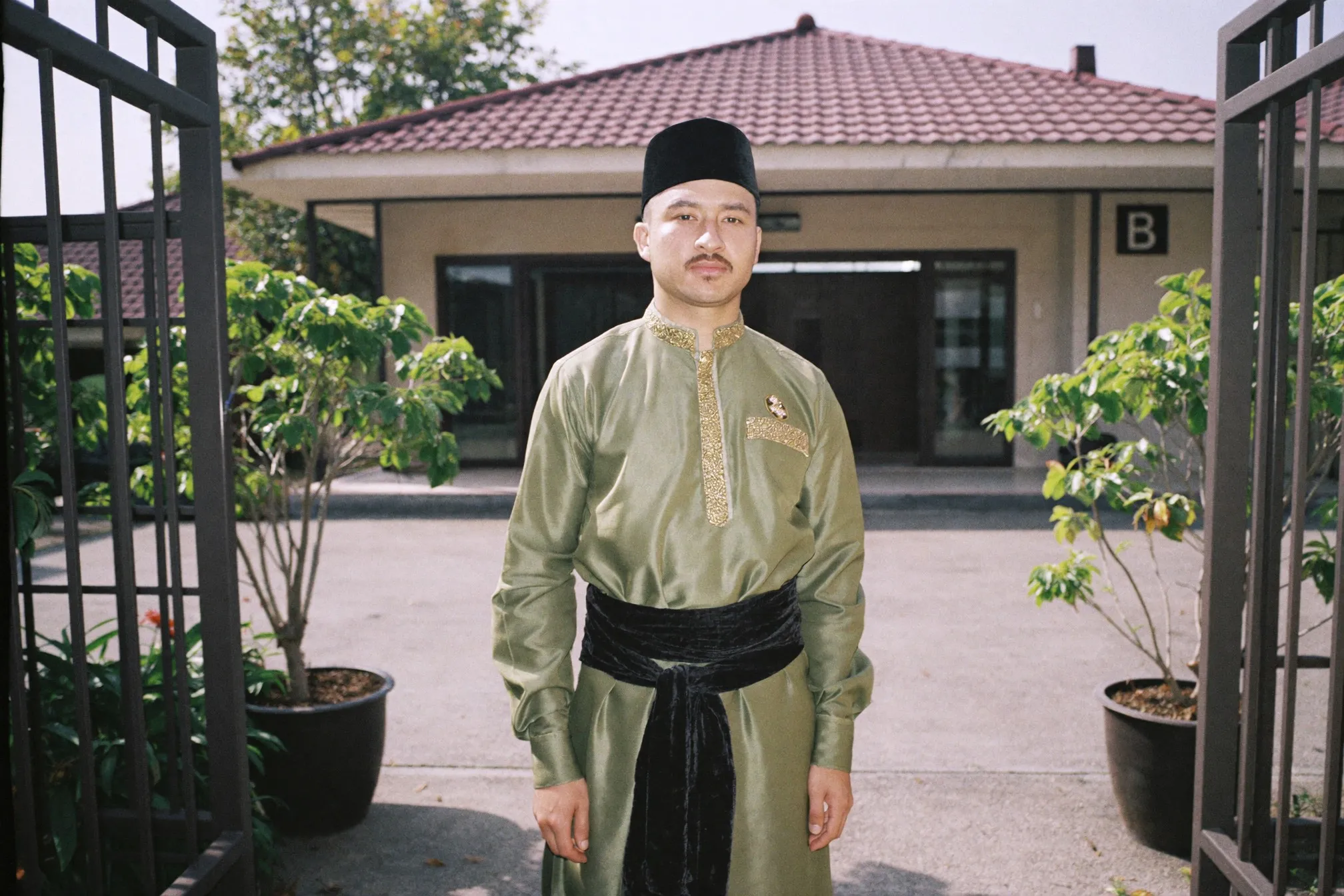 Man in a sage green baju melayu with a songket sampin and black songkok standing at the gate of a terrace house on Raya morning