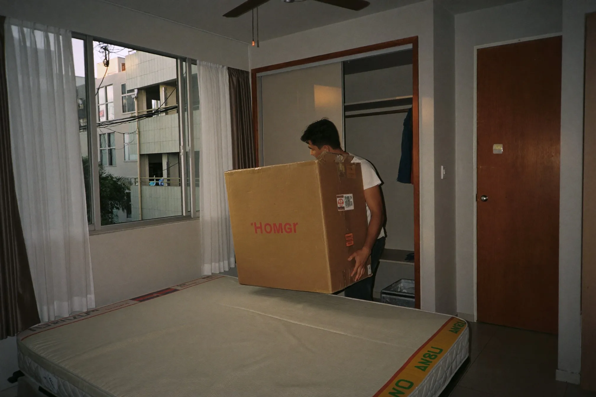 Young man carrying a moving box into a bright HDB bedroom with a bare mattress and an open window