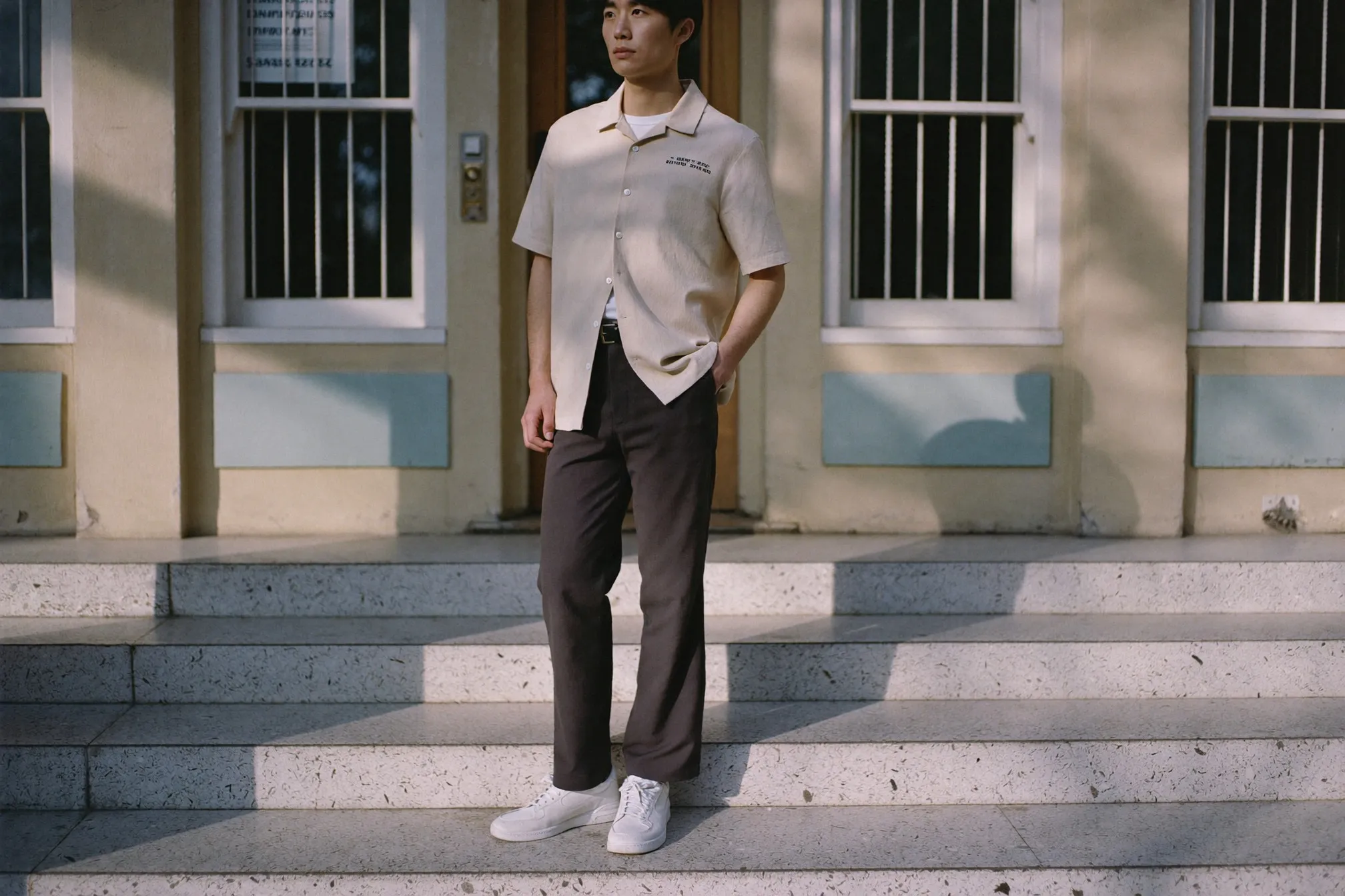 Man in a cream 1950s-style camp-collar shirt and modern dark trousers standing on the terrazzo steps of an old art deco shopfront in Kuala Lumpur