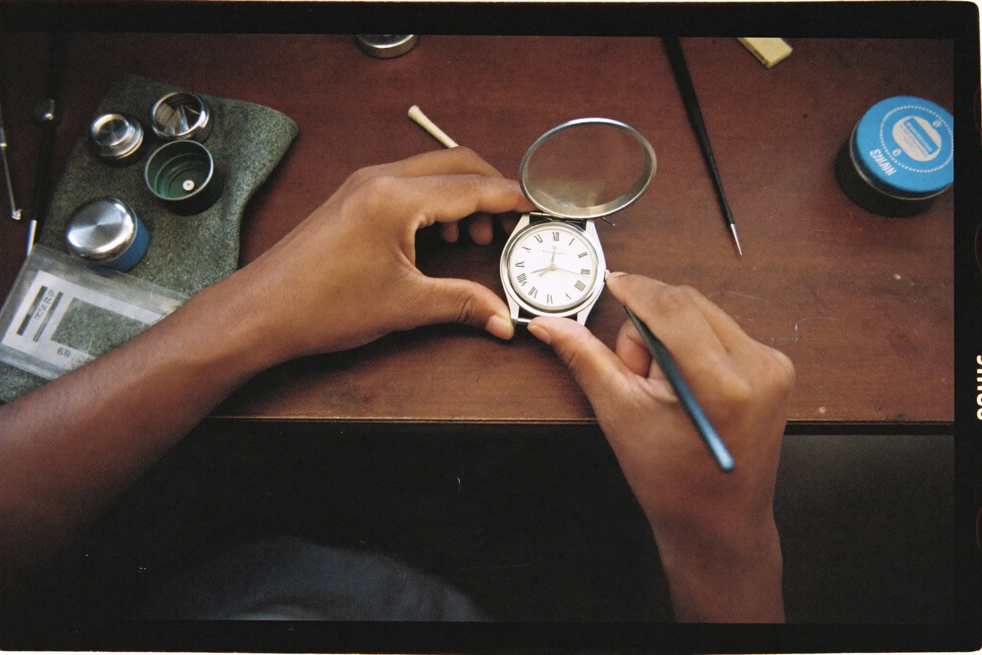 Watchmaker's hands with tweezers working on an opened plain steel watch case at a small repair shop bench