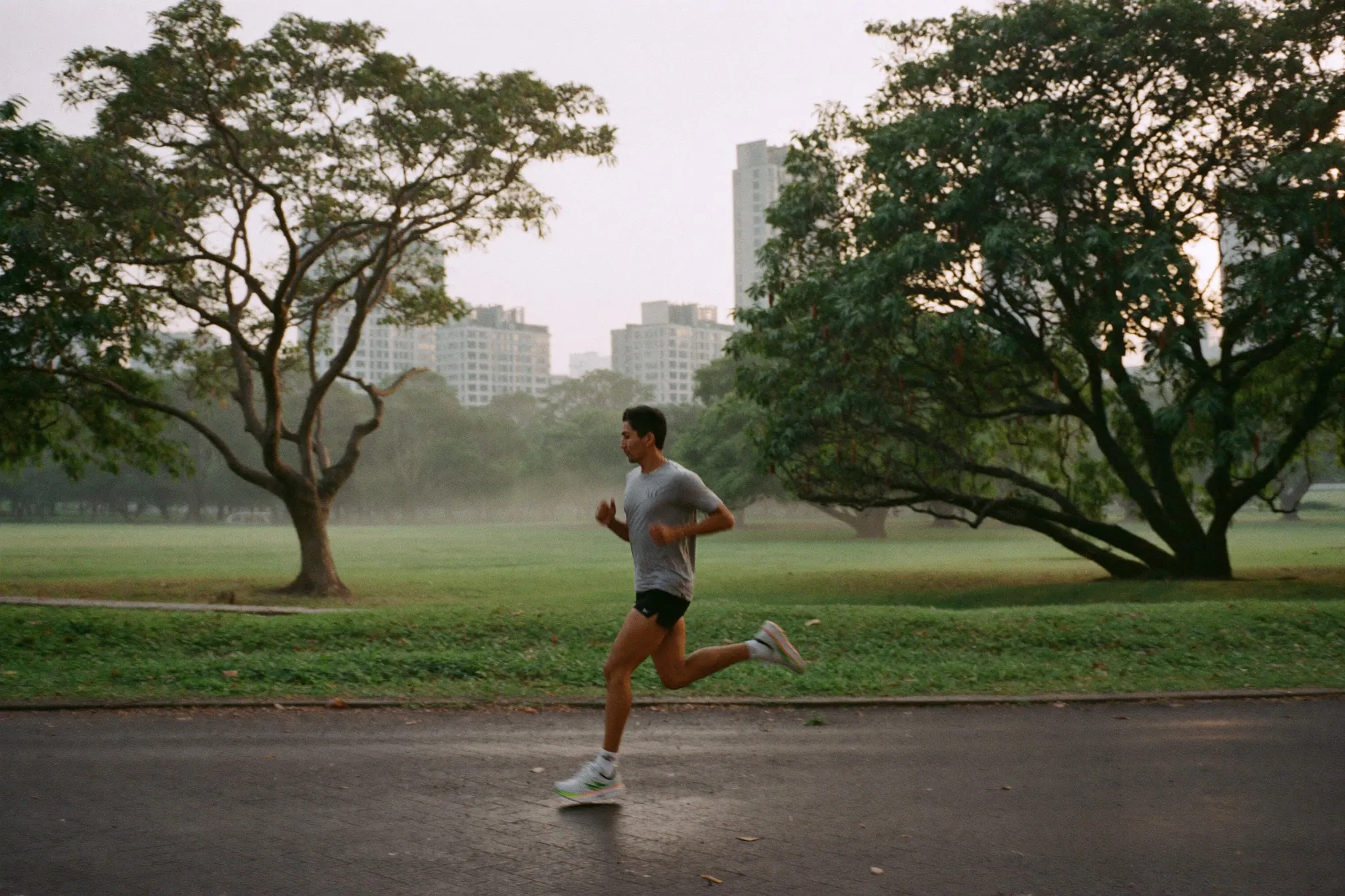 Malay man in his late twenties running along a park path in Kuala Lumpur just after sunrise with mist over the trees