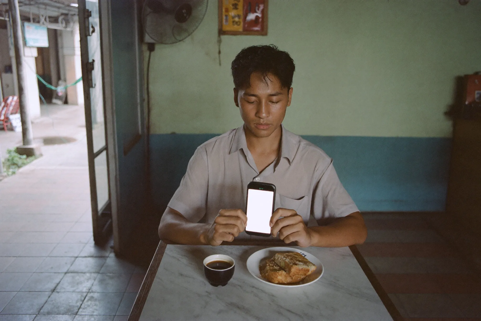 Young man checking his banking app at a kopitiam table in the morning with a coffee and a plate of toast