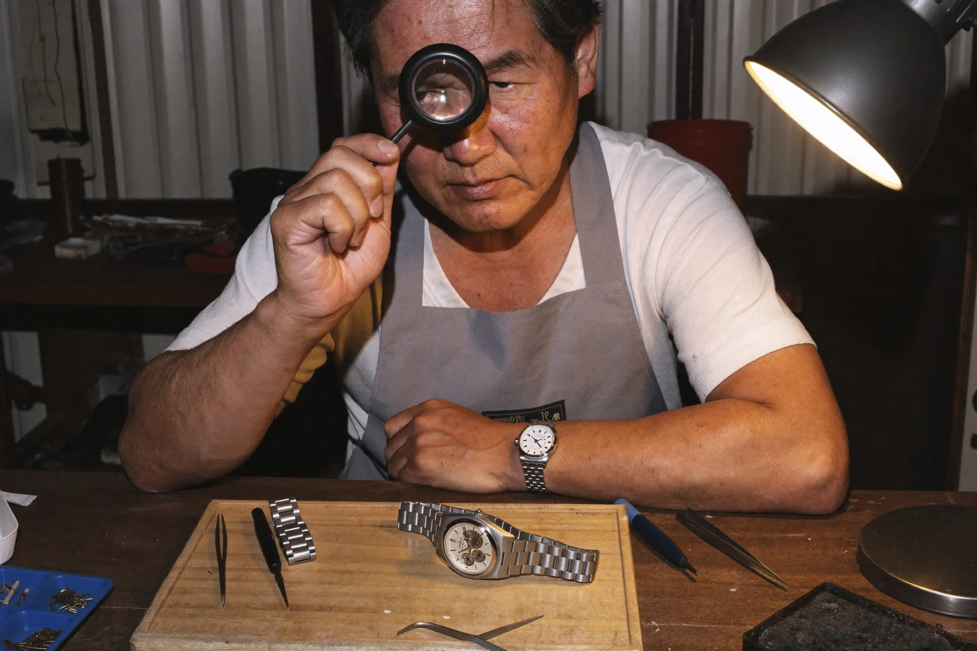 Watchmaker at a repair bench inspecting the exposed movement of an unbranded steel automatic watch through a loupe