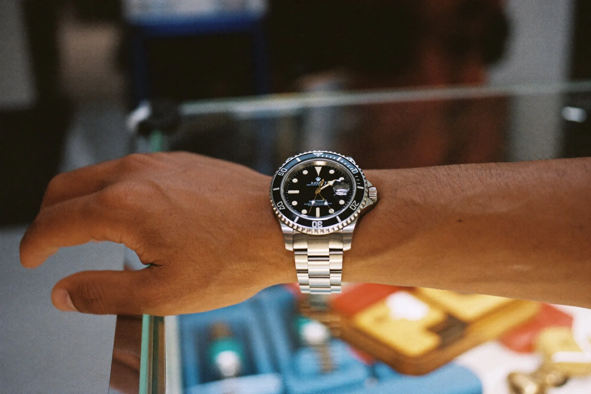 Man's wrist wearing a generic steel dive-style automatic watch, resting on the glass display counter of a mall watch shop in Kuala Lumpur