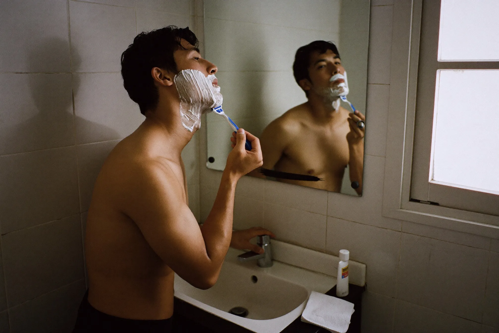 Man shaving his jawline in front of a bathroom mirror in a compact apartment, face covered in shaving foam