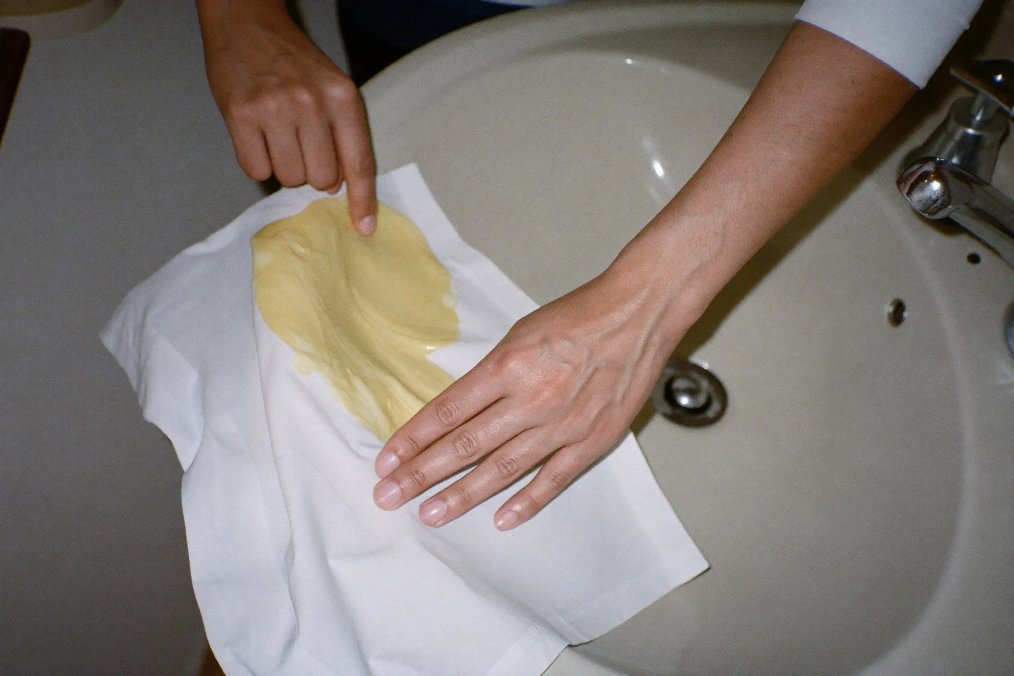 Close view of hands working stain treatment into the yellowed underarm of a white shirt at a bathroom sink