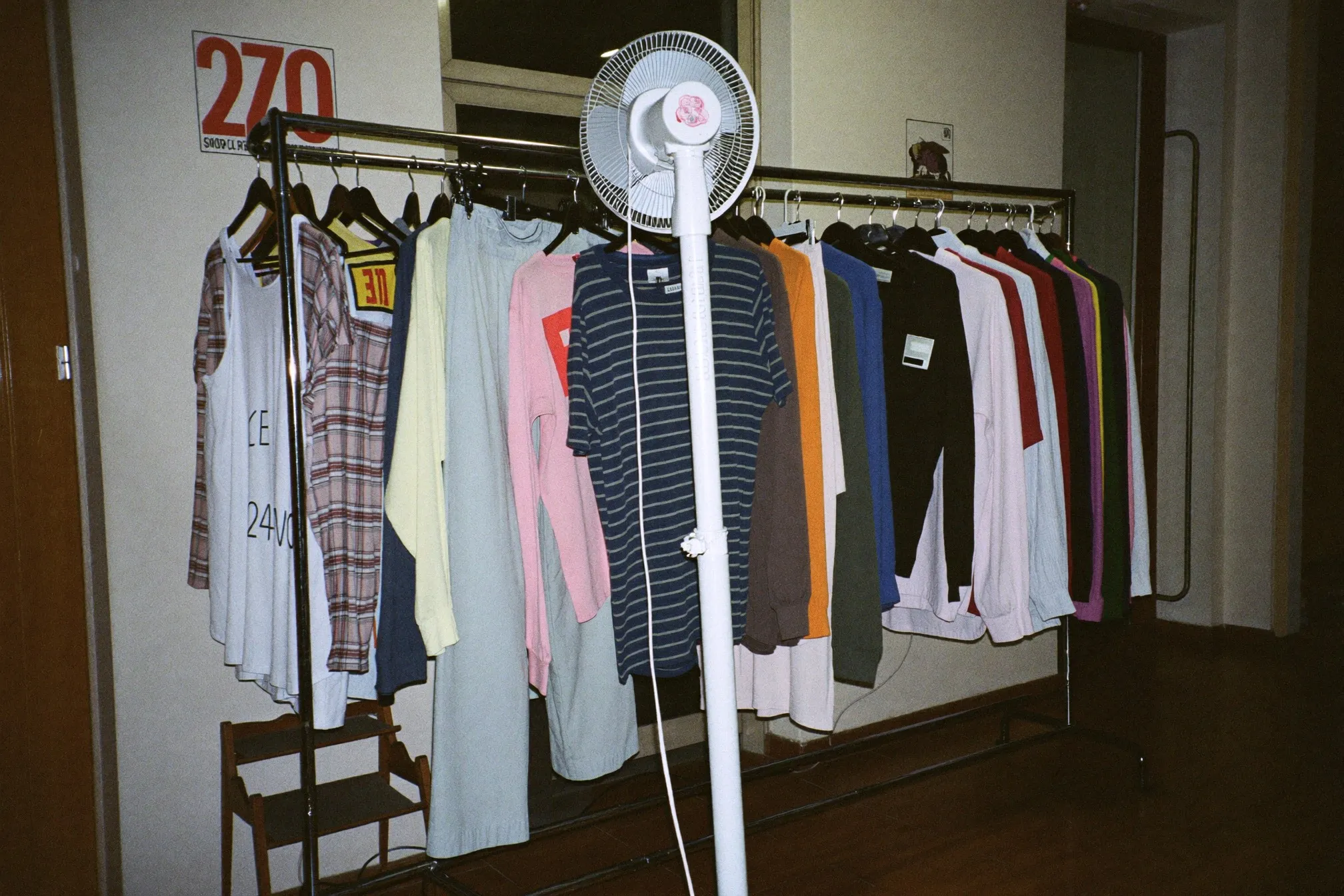 Shirts on hangers drying on a rack along an apartment corridor with an electric fan pointed at them