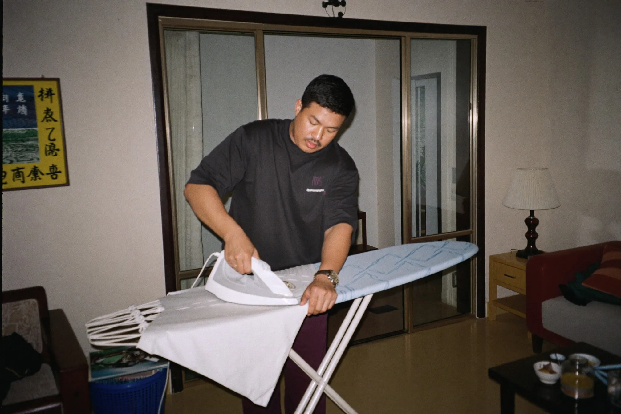 Man ironing a white cotton shirt on a folding board in the living room of an apartment