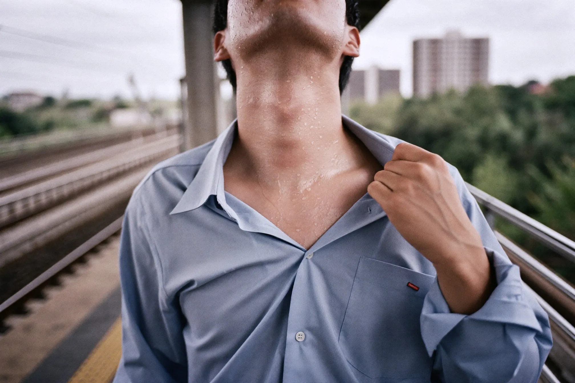 Man loosening his shirt collar at the neck while waiting on a humid open-air train platform