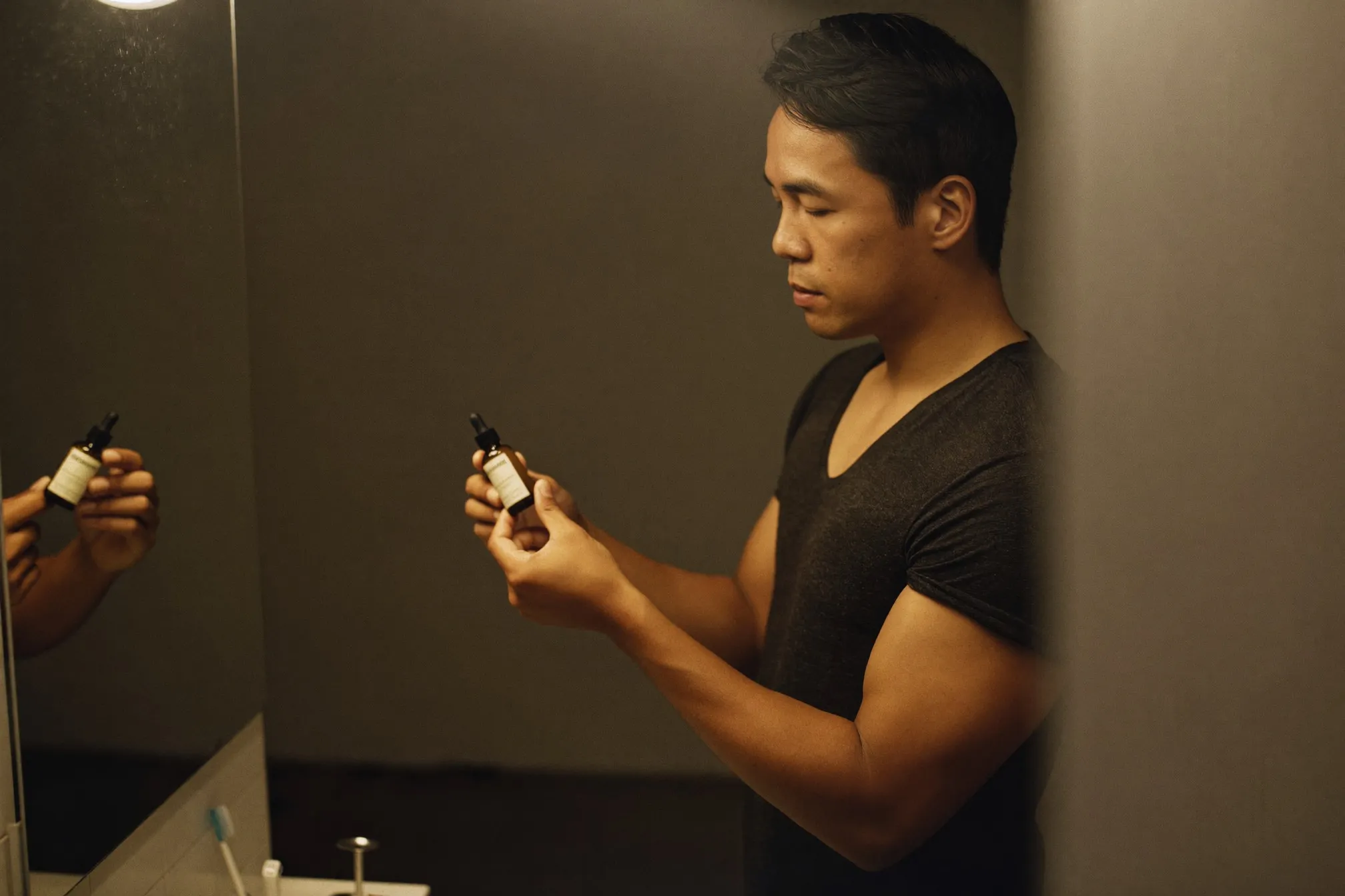 Man holding a small unbranded serum dropper bottle in front of a bathroom mirror at night