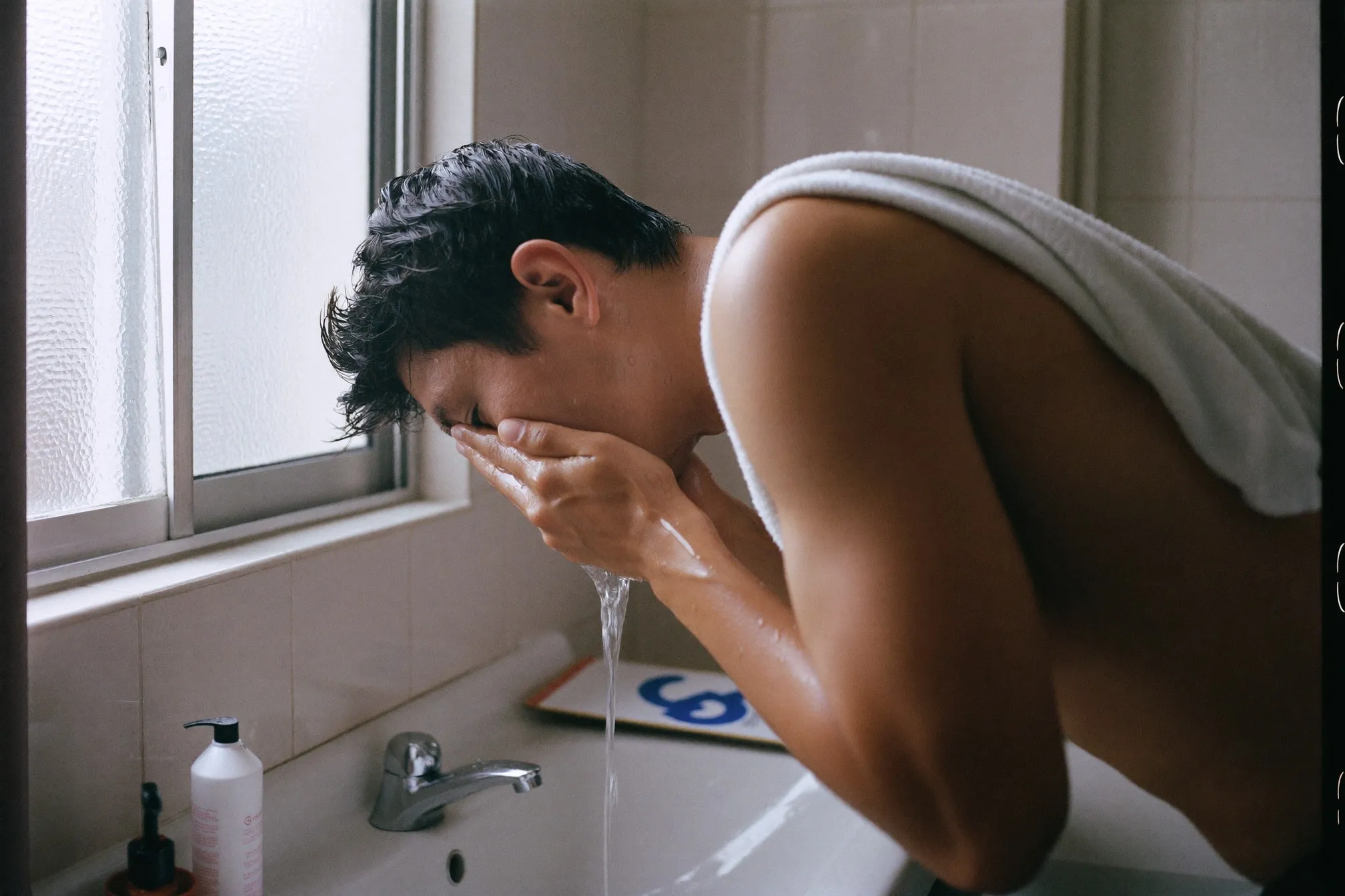 Man washing his face over a bathroom sink in the morning, towel over his shoulder