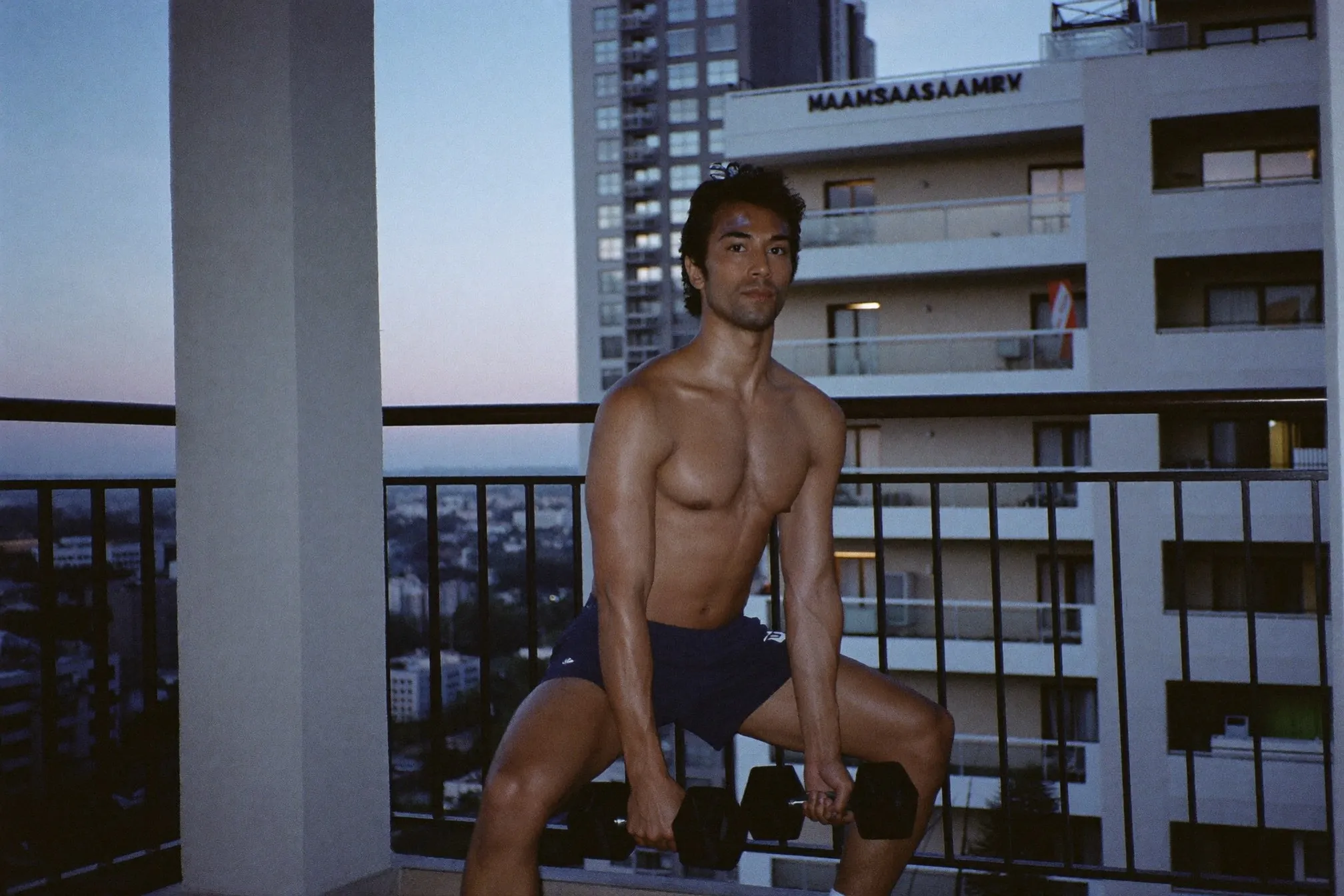 Man doing a goblet squat with a single dumbbell on the balcony of a Kuala Lumpur apartment at dusk