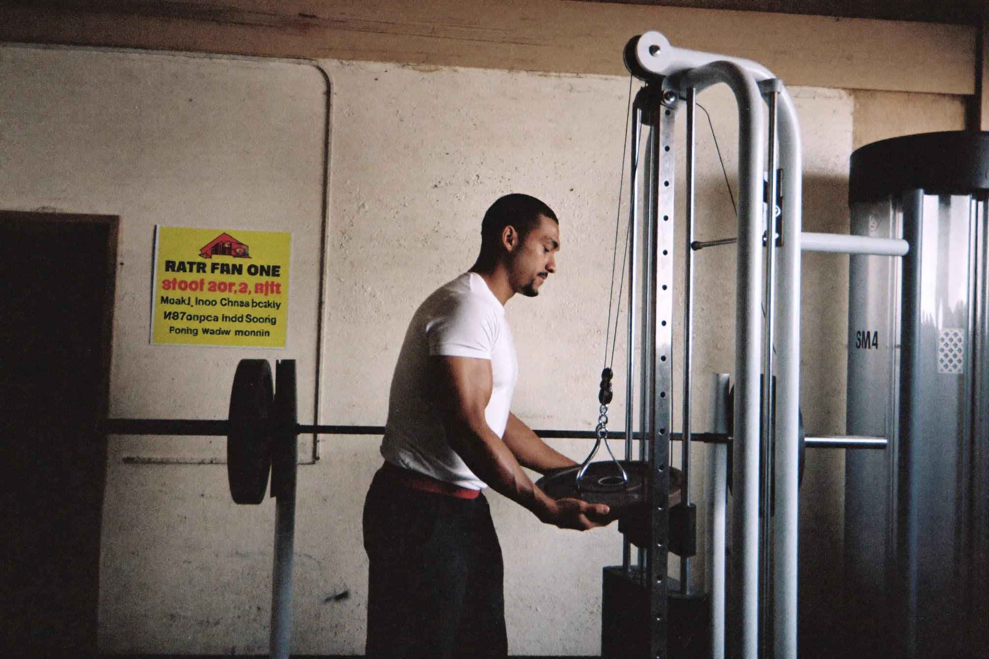 Man in a plain t-shirt loading a light plate onto a barbell at a no-frills community gym in the morning